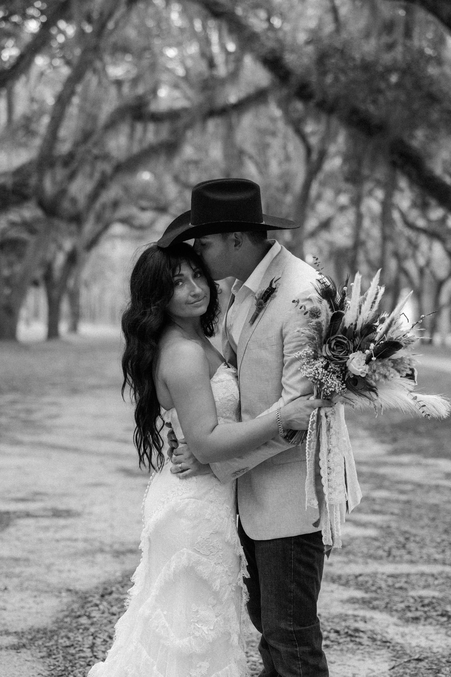 Black and white wedding portrait of a bride in a ruffled gown and a groom in a cowboy hat embracing beneath live oak trees.