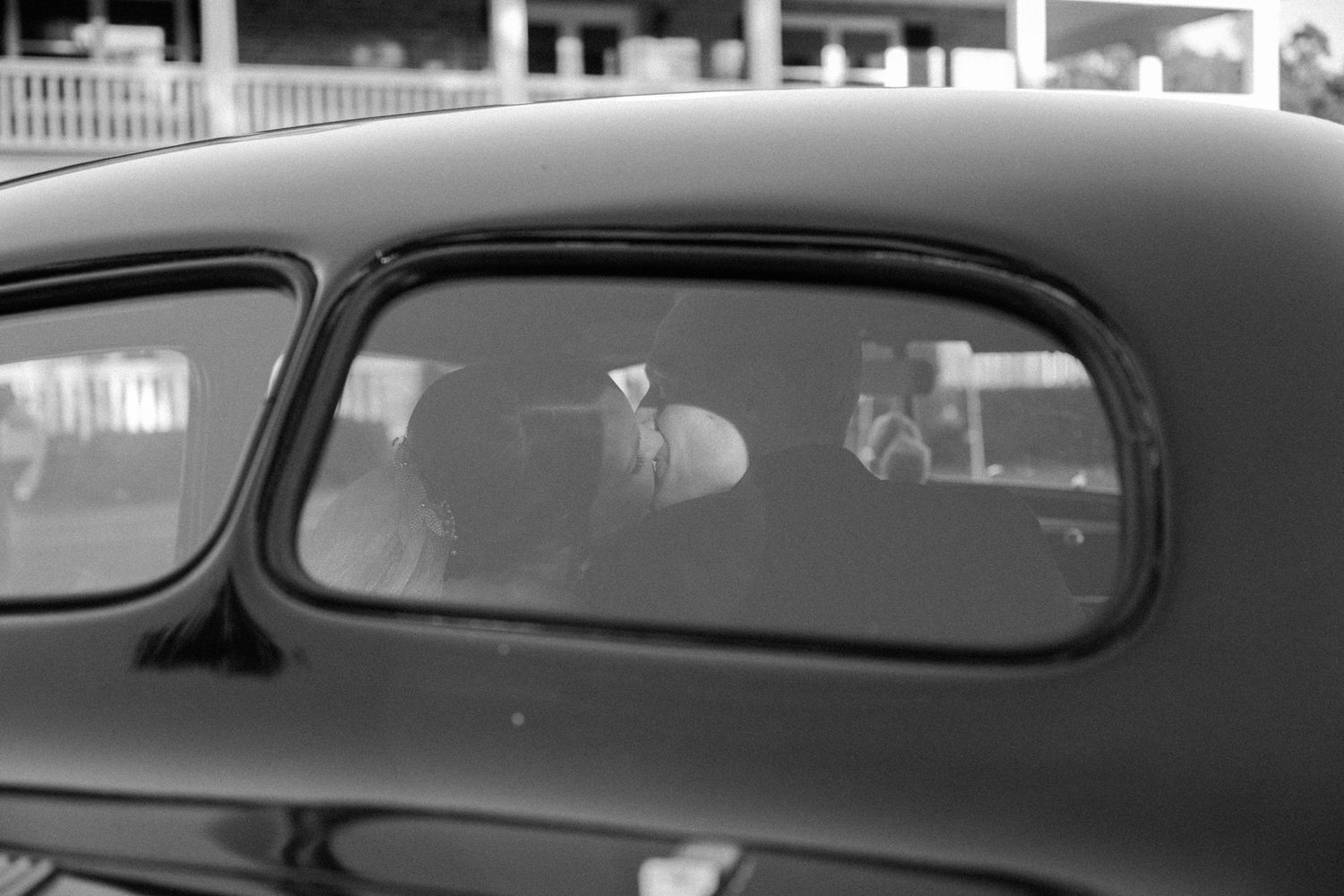 Black and white view through the window of a vintage car, with a couple faintly visible inside during a wedding day.