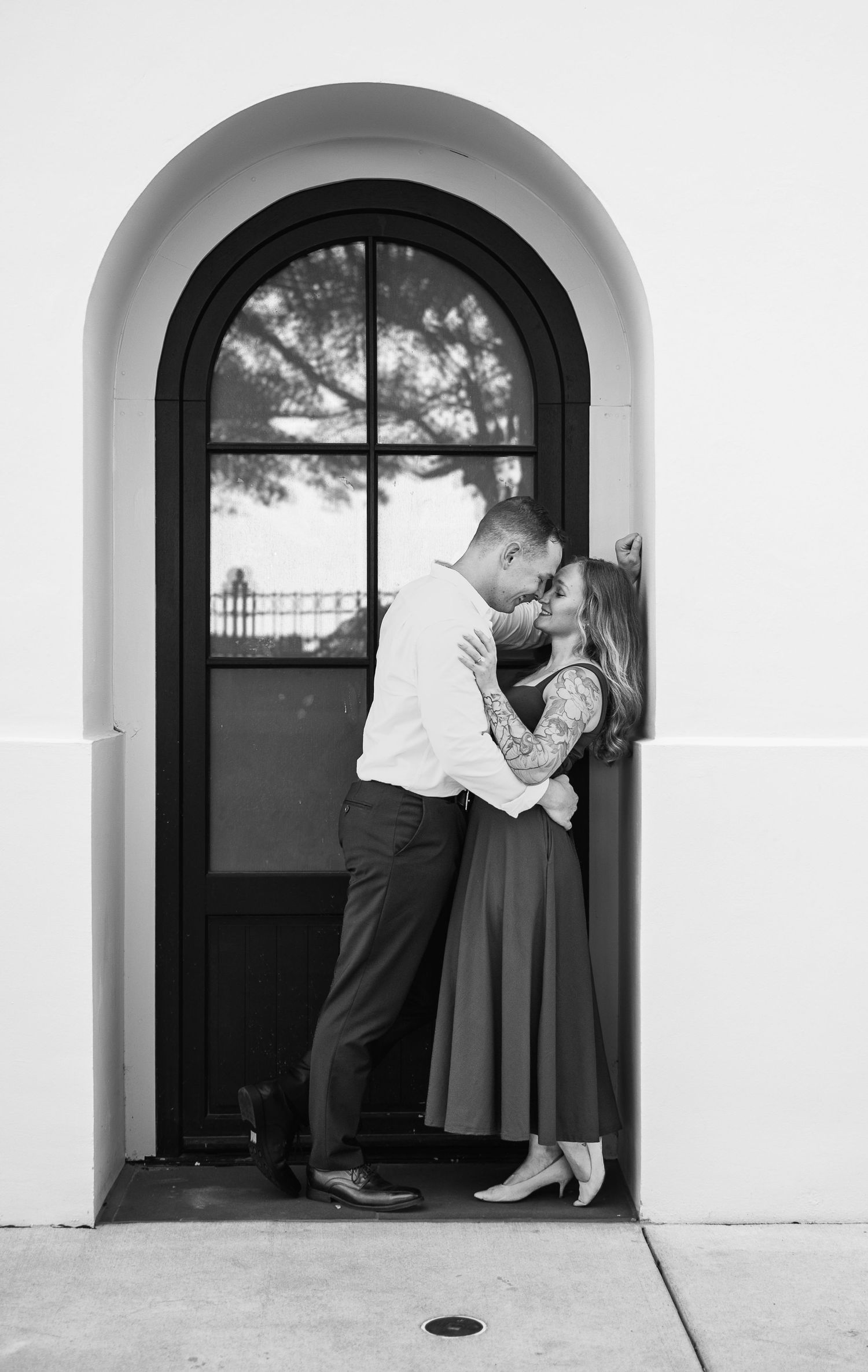 Black and white engagement photo of a couple nearly kissing in an arched doorway with a tall paned window.