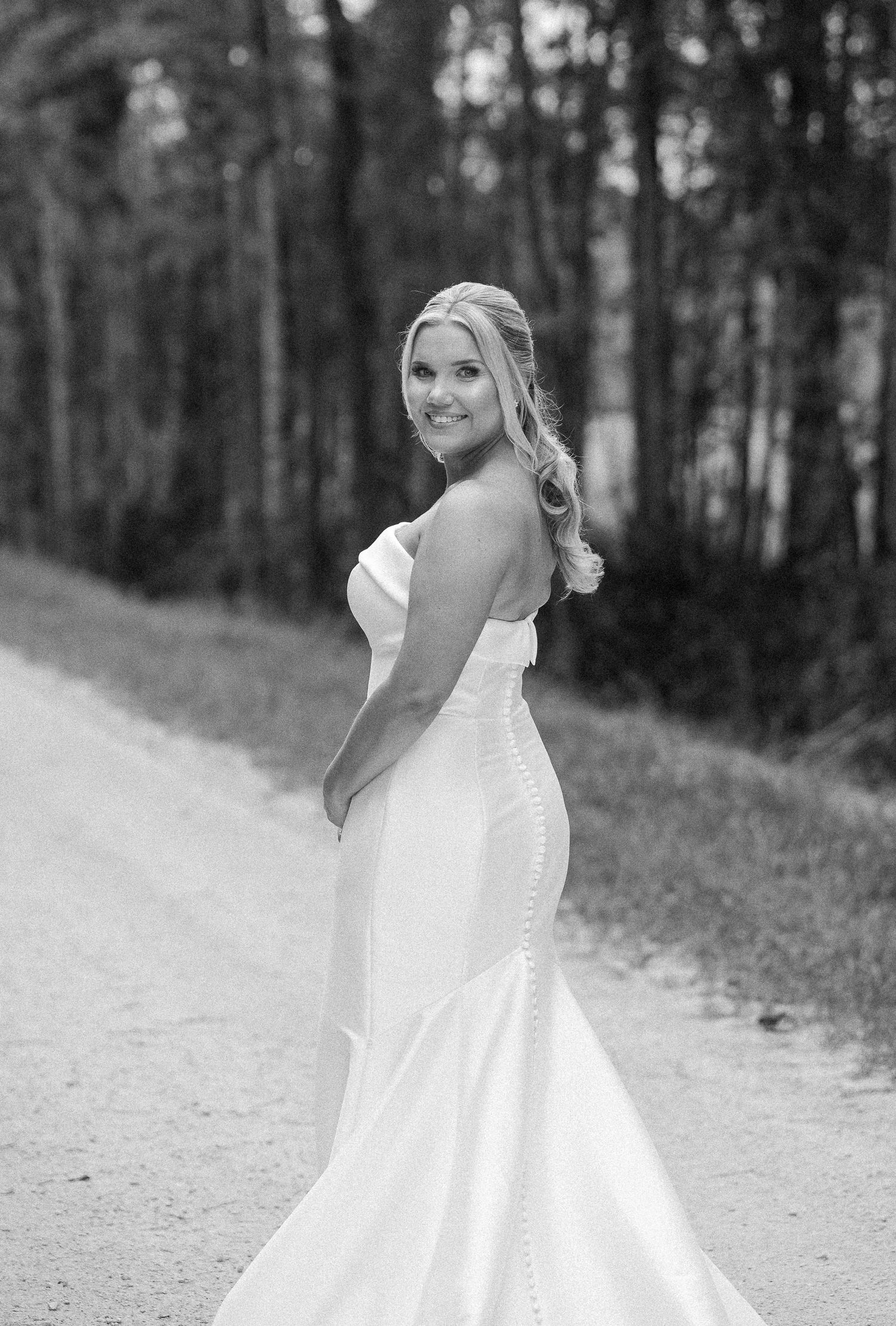 Black and white portrait of a smiling bride in a strapless satin wedding gown glancing over her shoulder on a country road.