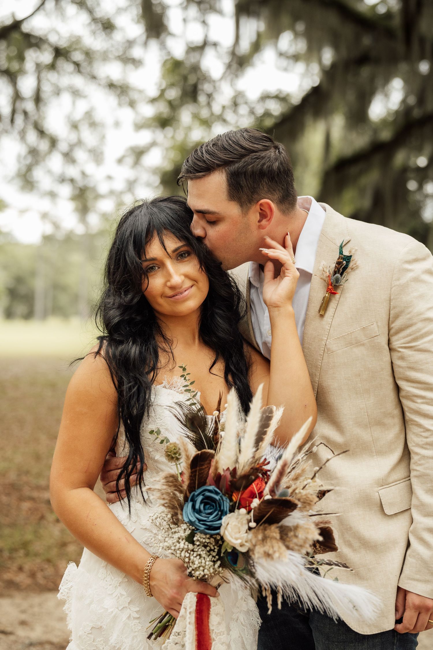 Smiling bride in a lace wedding dress holds a pampas grass bouquet with a blue rose while her groom kisses her cheek outdoors.
