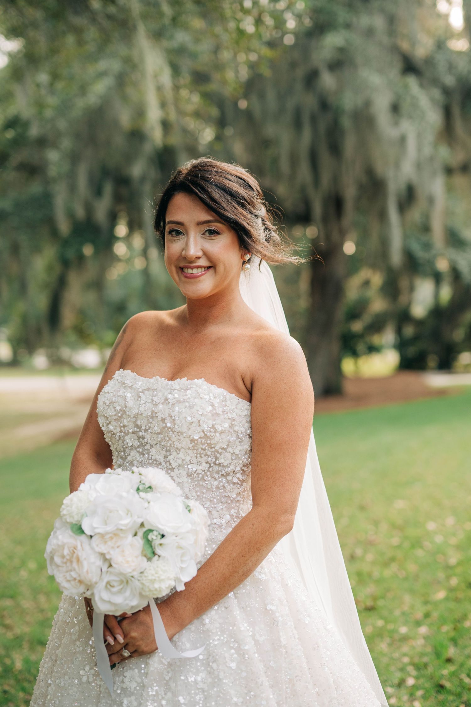 Smiling bride in a strapless beaded gown and veil holding a white peony bouquet among moss-draped trees.