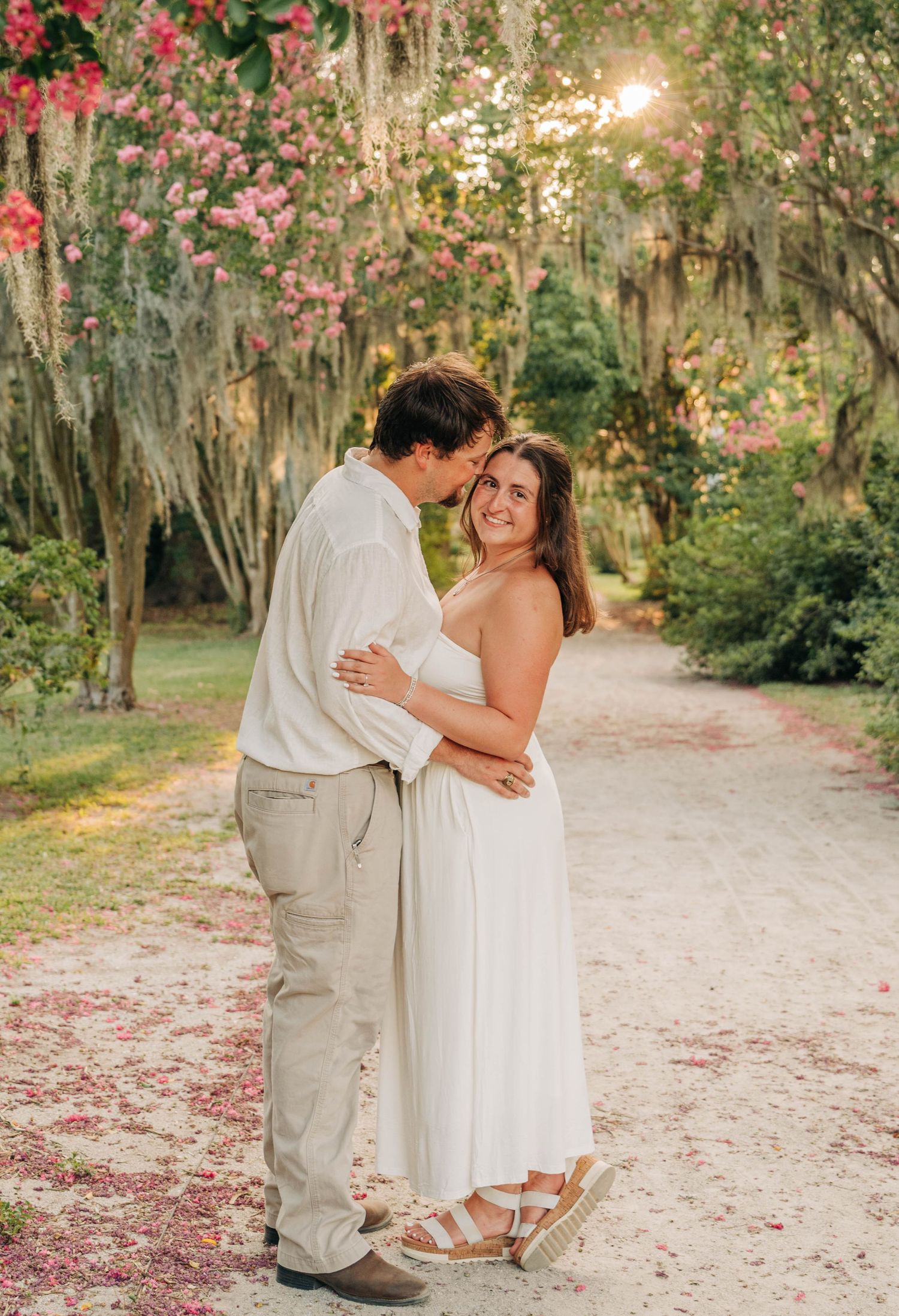 Engaged couple embraces on a sandy path beneath oak trees draped in Spanish moss and pink blossoms at golden hour.