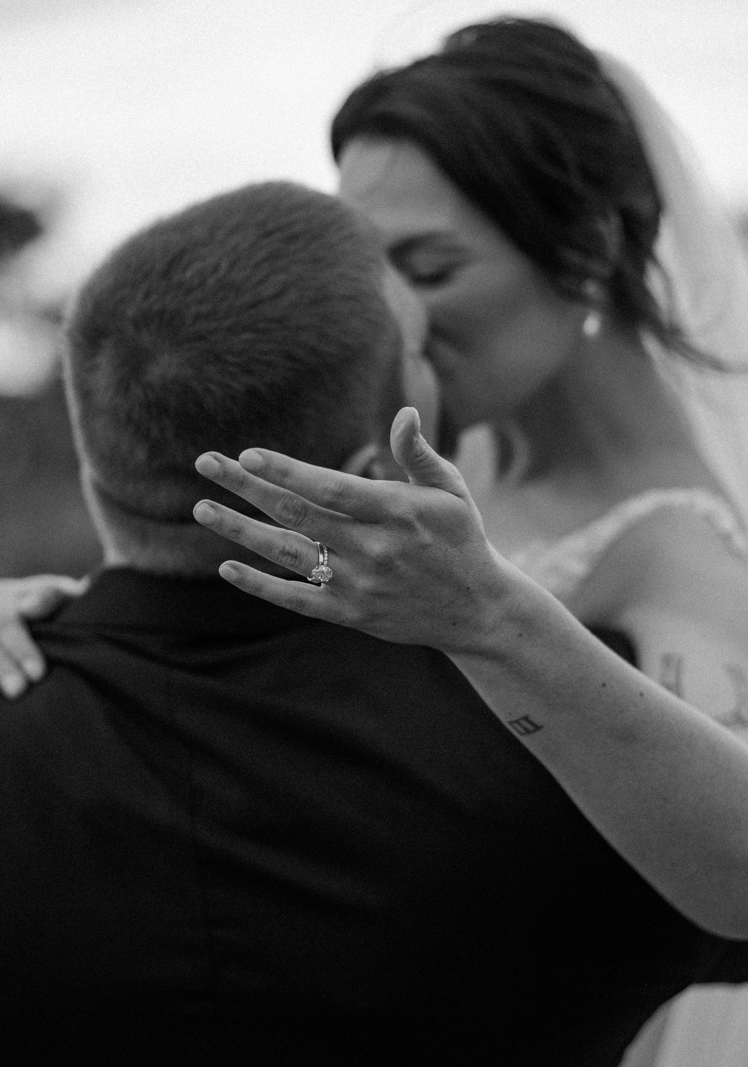Black and white photo of a bride and groom kissing as her hand with a sparkling engagement ring rests on his shoulder.