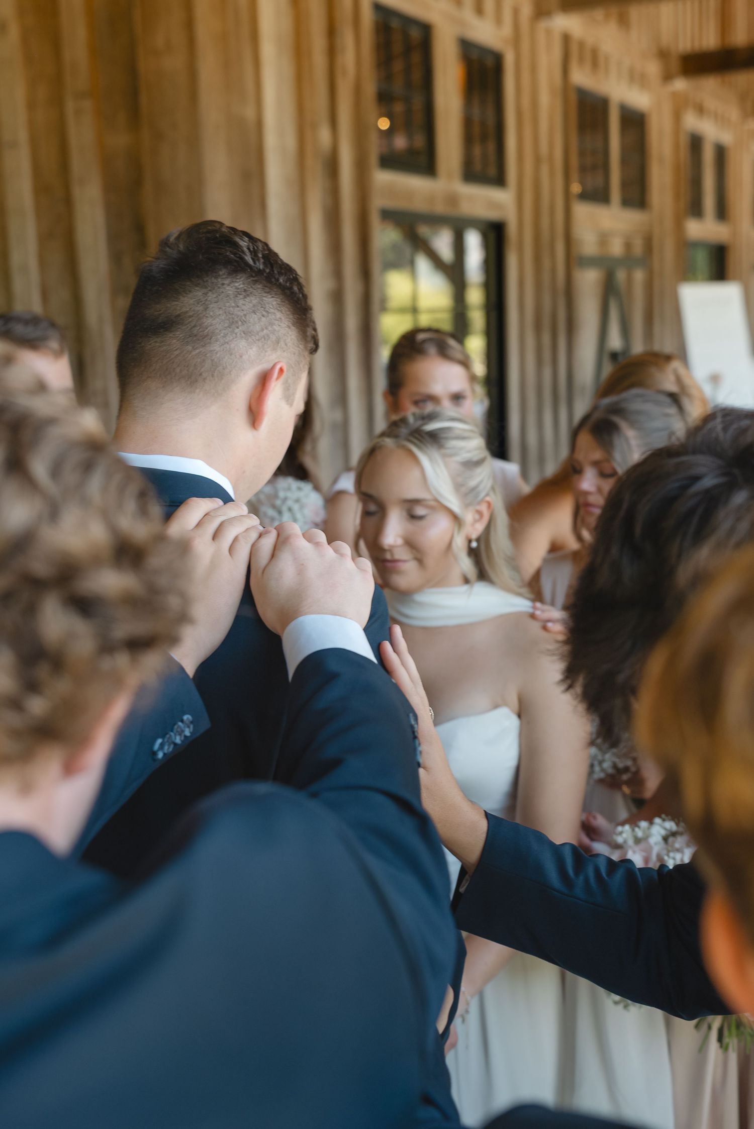Groom and bride stand close as family and bridal party members lay hands on their shoulders in prayer inside a rustic wooden venue.
