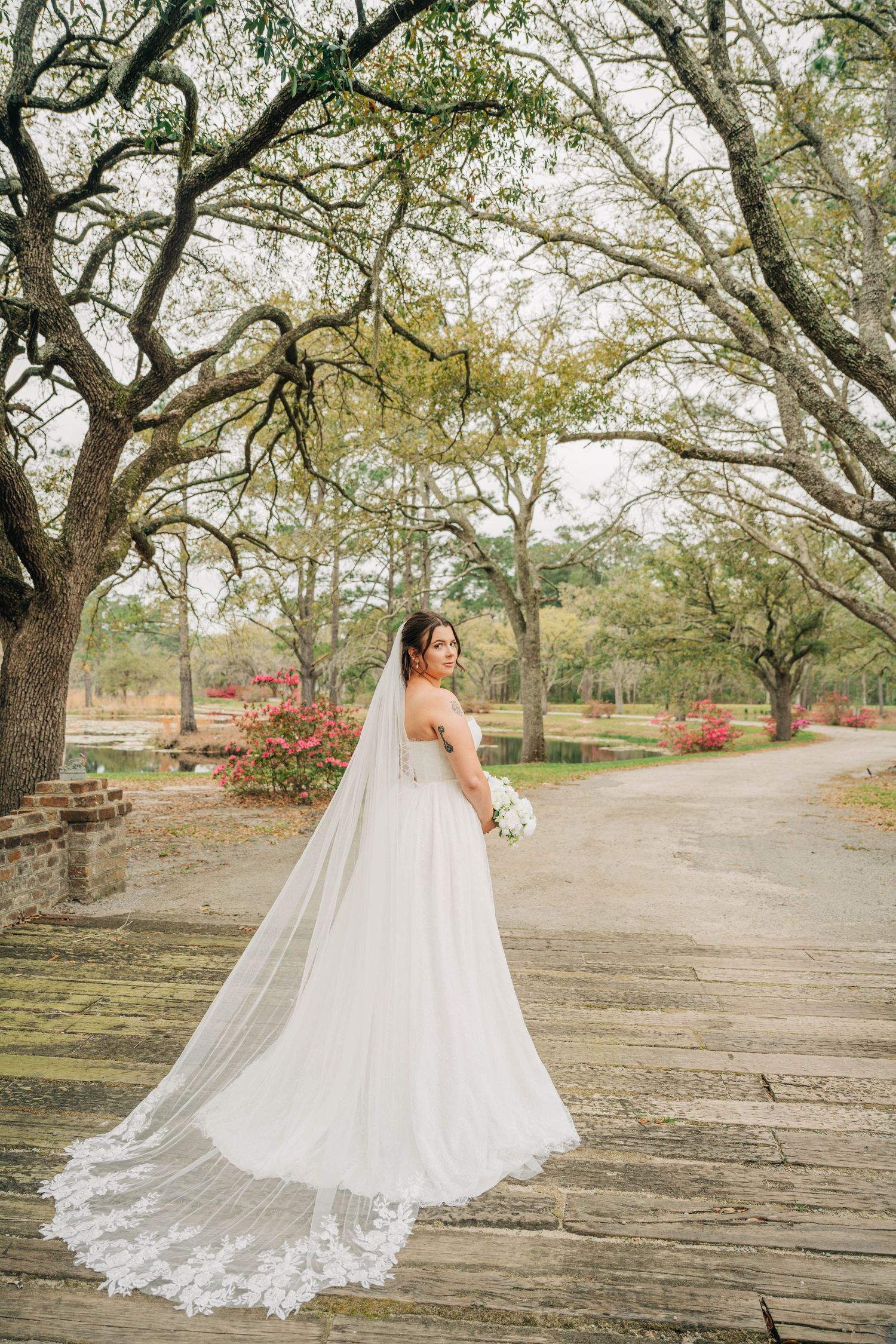Bride in a tulle gown with a long lace-trimmed veil stands on a wooden boardwalk under oak trees in a garden.