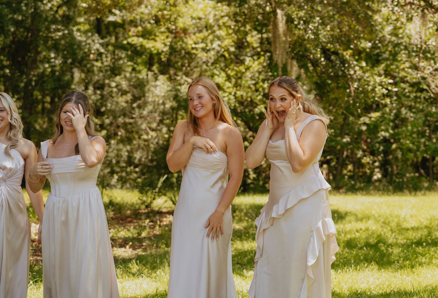 Three bridesmaids in flowing champagne dresses cover their faces and tear up with emotion while standing on a sunlit grassy lawn.