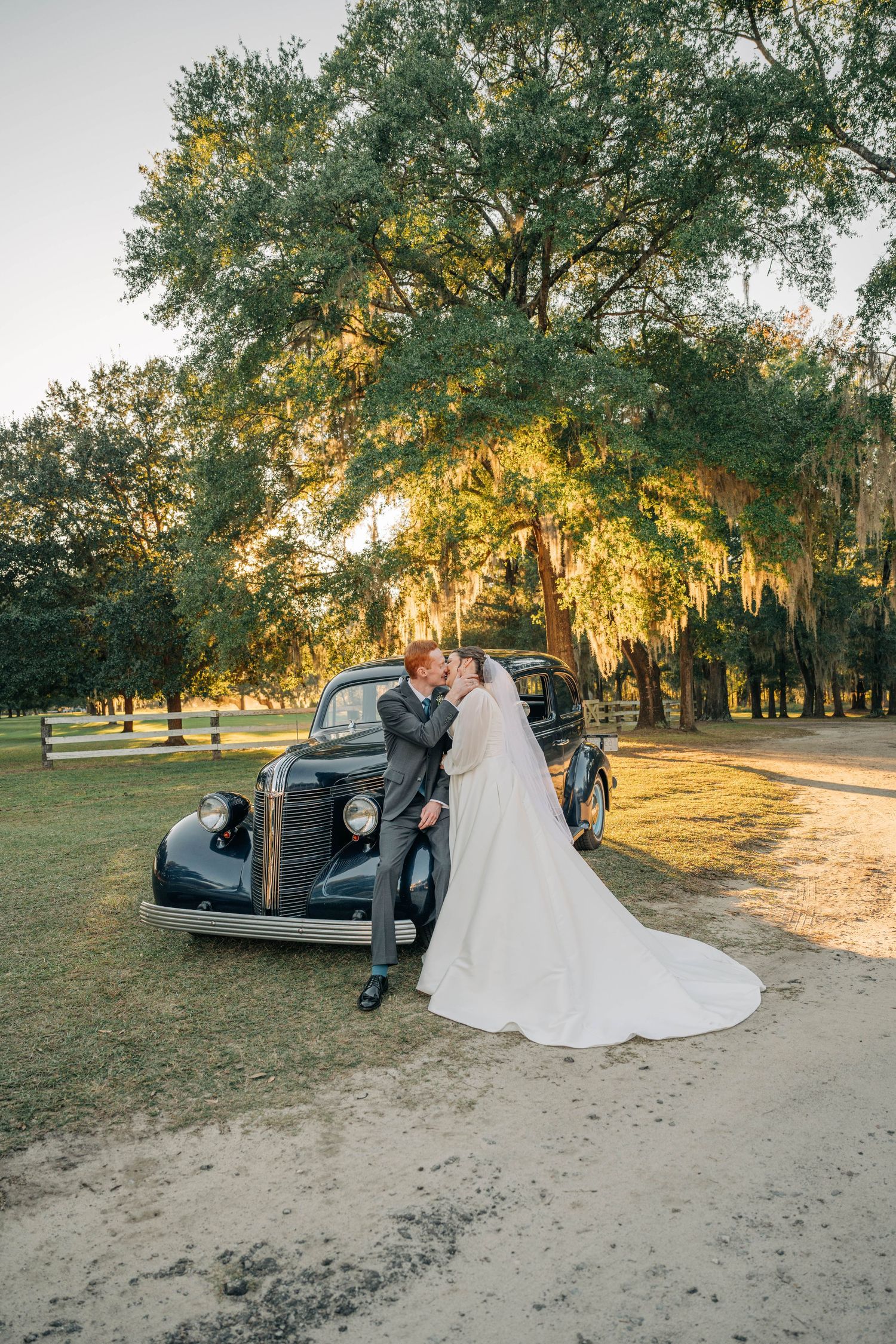 Bride and groom kiss beside a vintage black car under large oak trees draped in Spanish moss at golden hour.