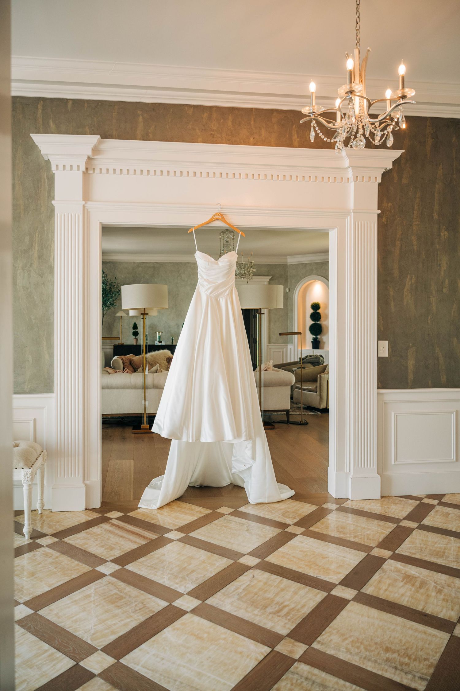 White wedding dress hanging in an elegant white archway above a geometric tiled floor with a chandelier overhead.