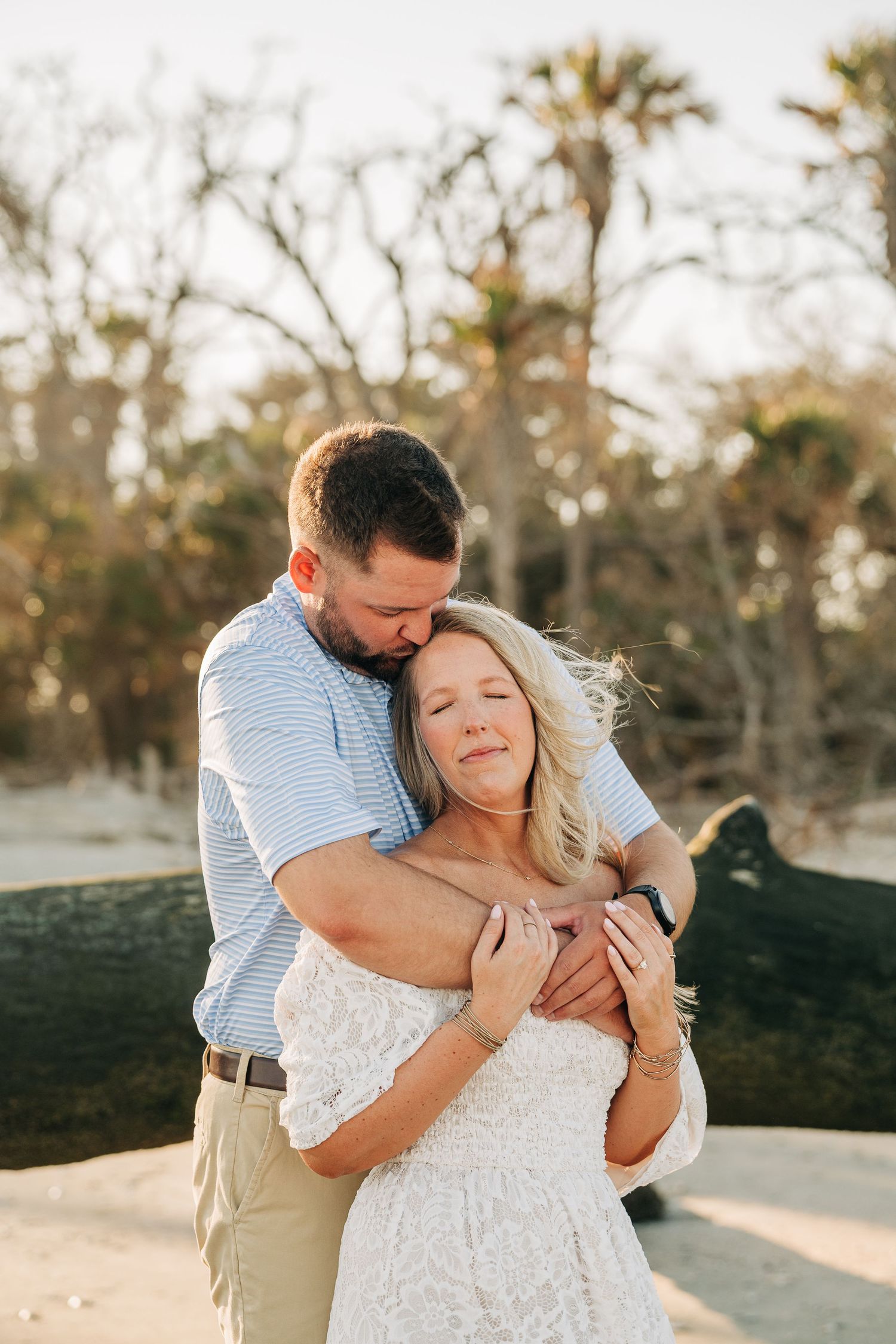 Man hugging his fiancée from behind on a sunny beach, her eyes closed in a peaceful moment during an engagement session.
