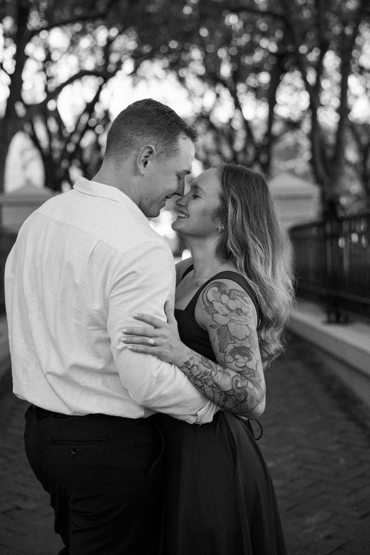 Black and white engagement photo of a smiling couple embracing nose to nose beneath trees, the woman with a floral sleeve tattoo.
