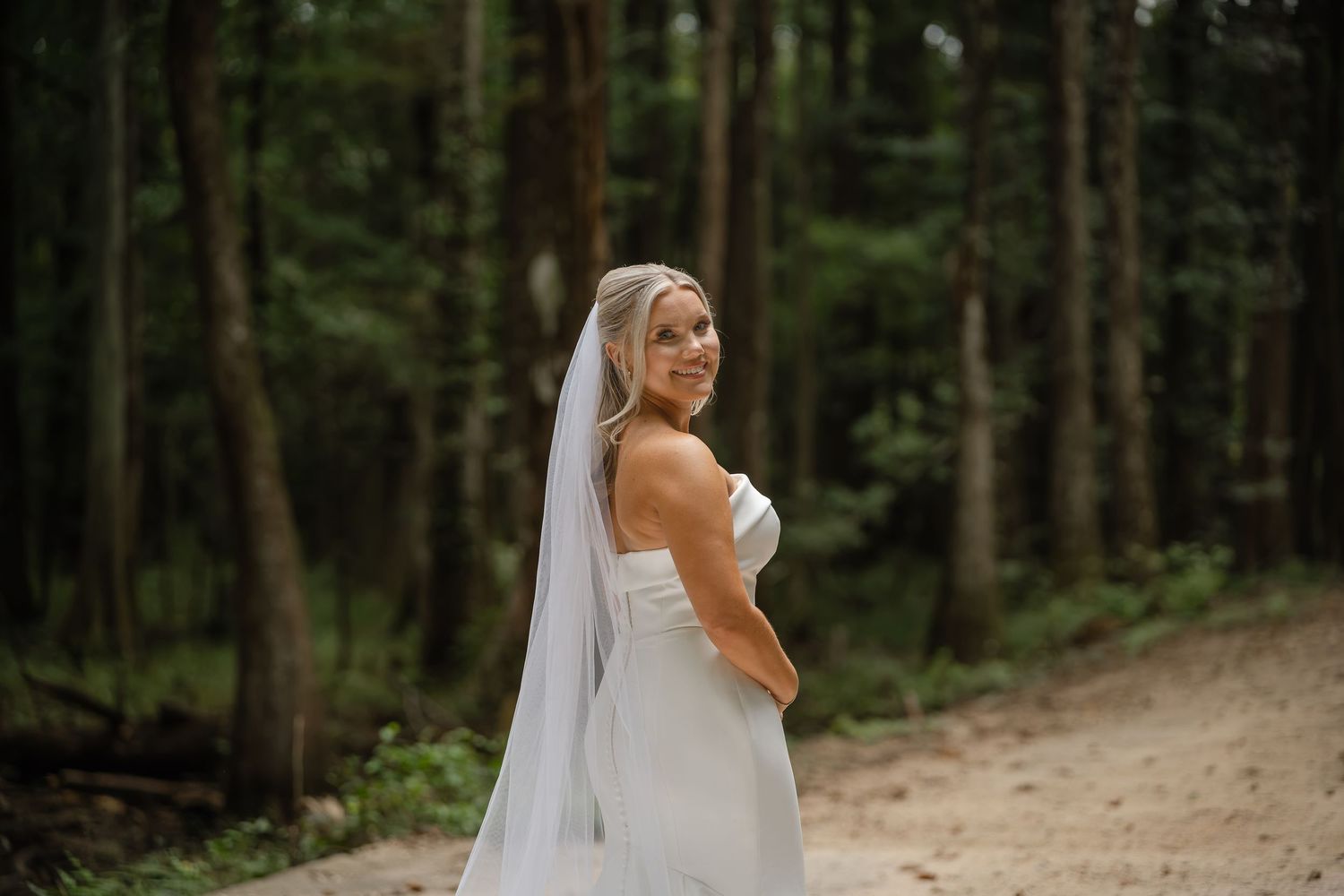 Bride wearing a long cathedral veil and strapless gown smiles over her shoulder on a path through a forest.