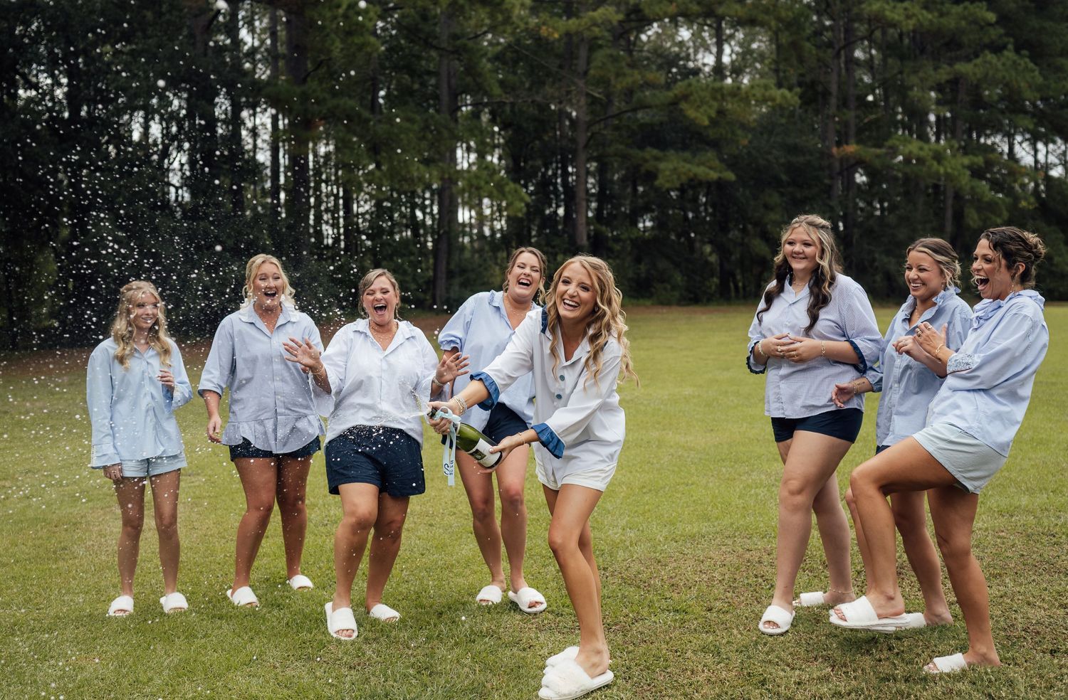 Bridesmaids in matching button-up shirts laugh and spray champagne on a grassy lawn surrounded by tall pine trees.