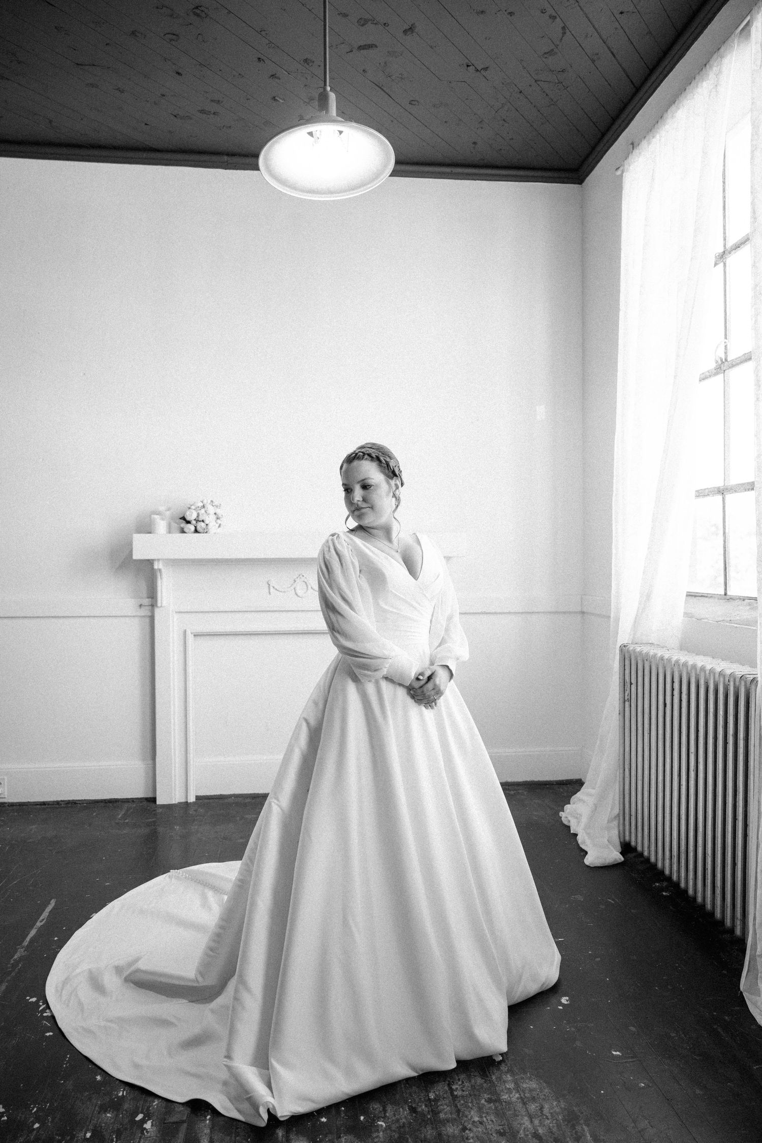 Black and white portrait of a bride in a long-sleeved satin gown with a cathedral train standing in a bright room with a vintage radiator.