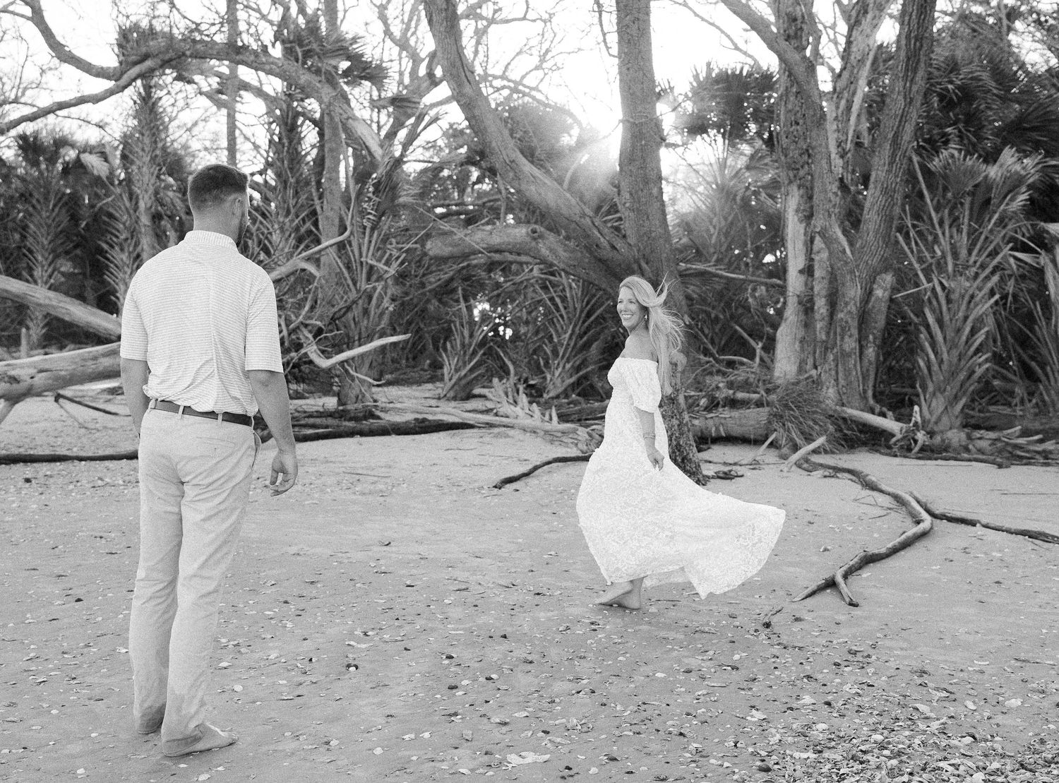 Black and white photo of a woman in a flowing white dress twirling toward a man on a beach among driftwood and palms.