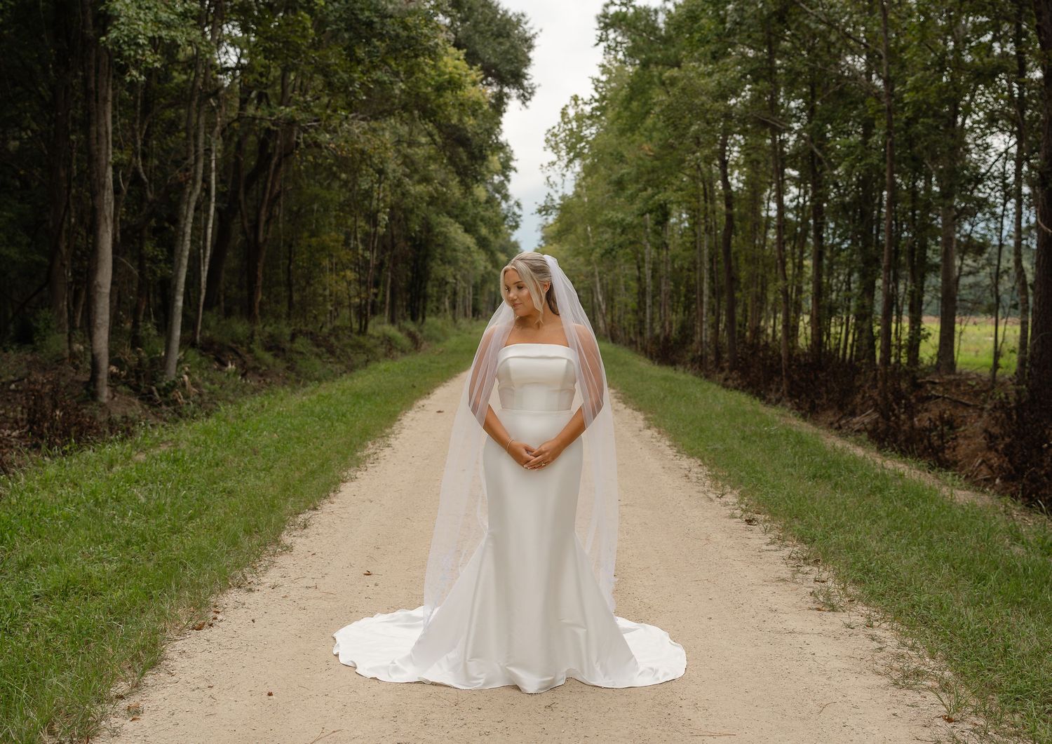 Bride in a strapless white gown and flowing veil poses on a gravel road surrounded by tall trees and green grass.