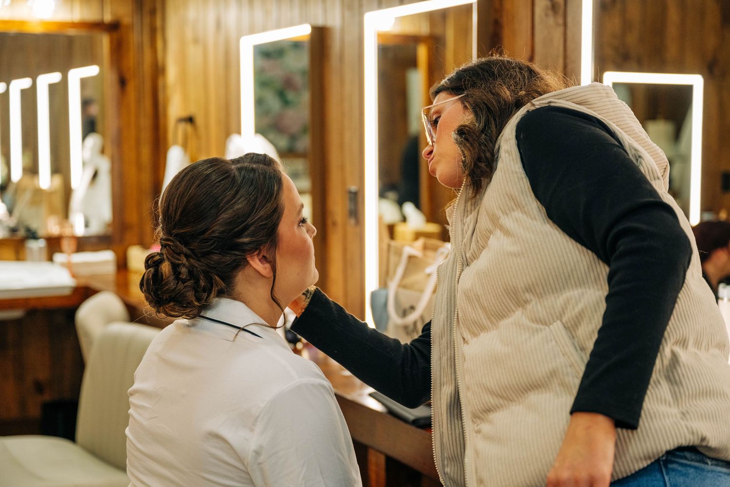 A makeup artist applies bridal makeup to a bride with an elegant updo in a warm, wood-paneled getting-ready room.