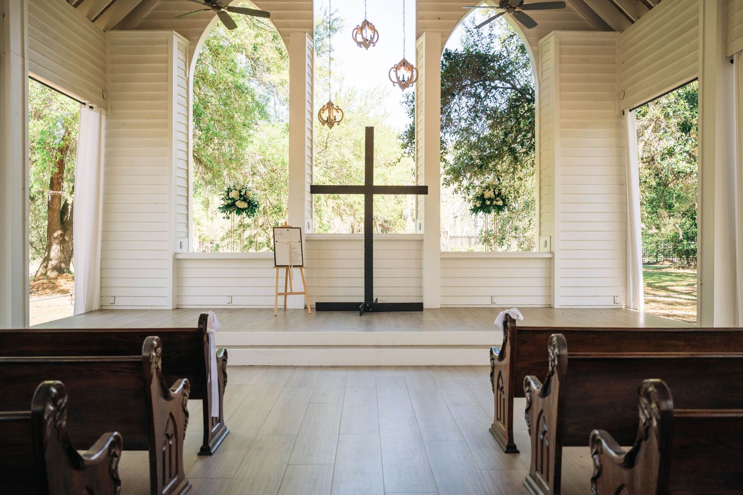 Interior of a white chapel with wooden pews, a central black cross and arched windows overlooking green trees.