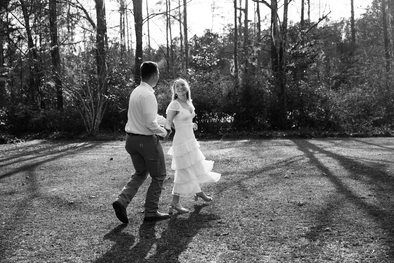 Black and white photo of a couple dancing outdoors on a sunlit lawn with trees and long shadows in the background.