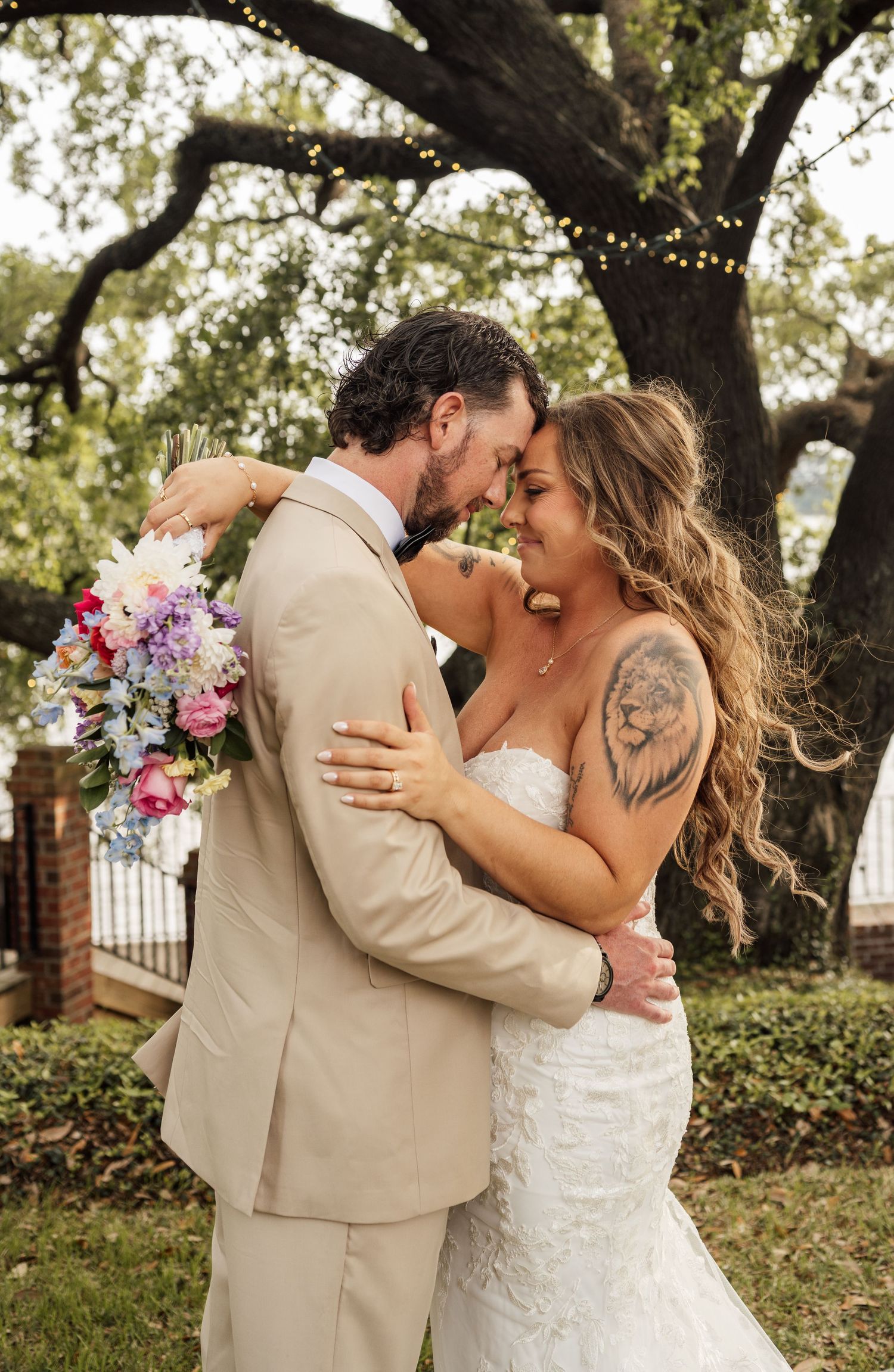 Bride in a lace gown and groom in a tan suit embrace under a mossy oak tree, holding a colorful wildflower bouquet.