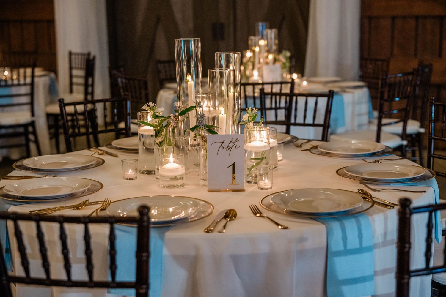 Elegant wedding reception table set with white linens, plates, candles and a numbered table sign amid warm glowing light.