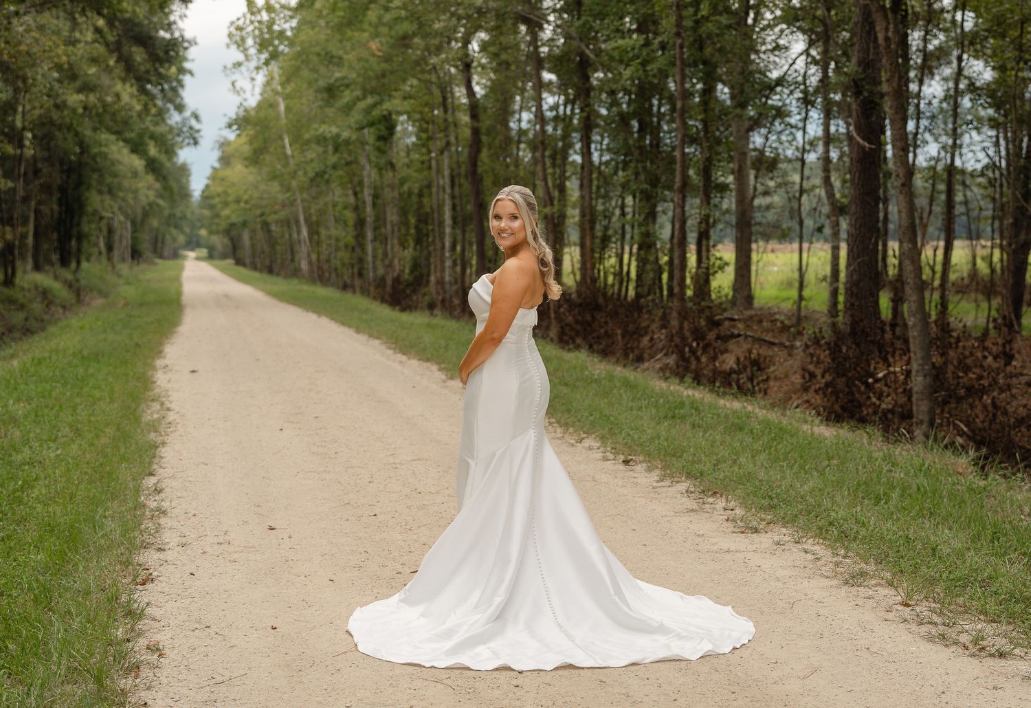Bride in a white mermaid wedding gown with a long train turns to smile on a gravel road lined with tall trees.