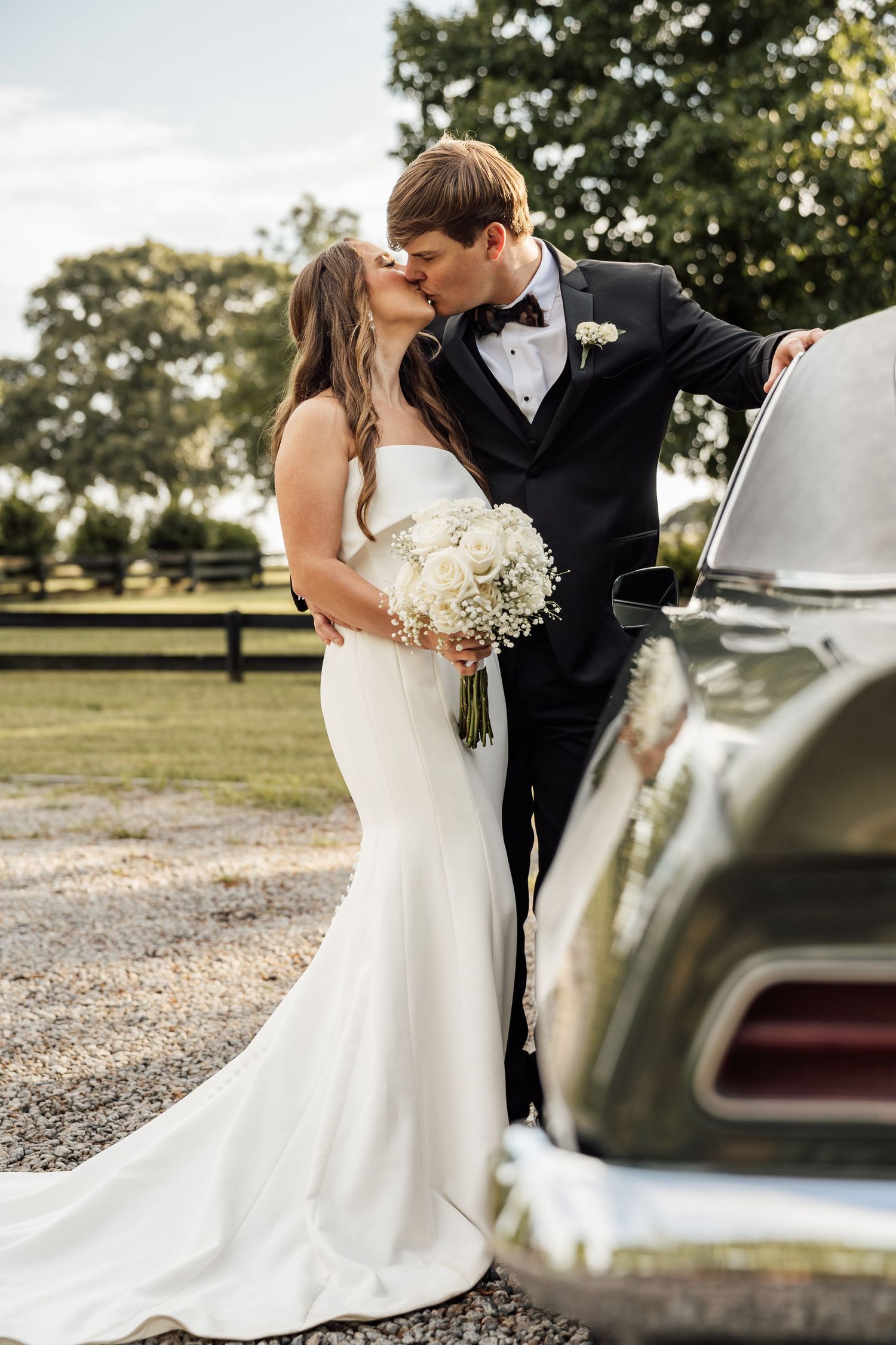 Bride holding a white bouquet kisses the groom in a black tuxedo beside a classic vintage car at an outdoor venue.