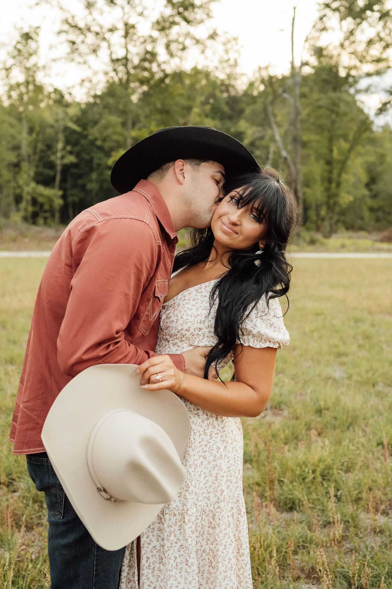 Man in a black cowboy hat kisses the cheek of a smiling woman in a floral dress while holding a cream hat in a green field.