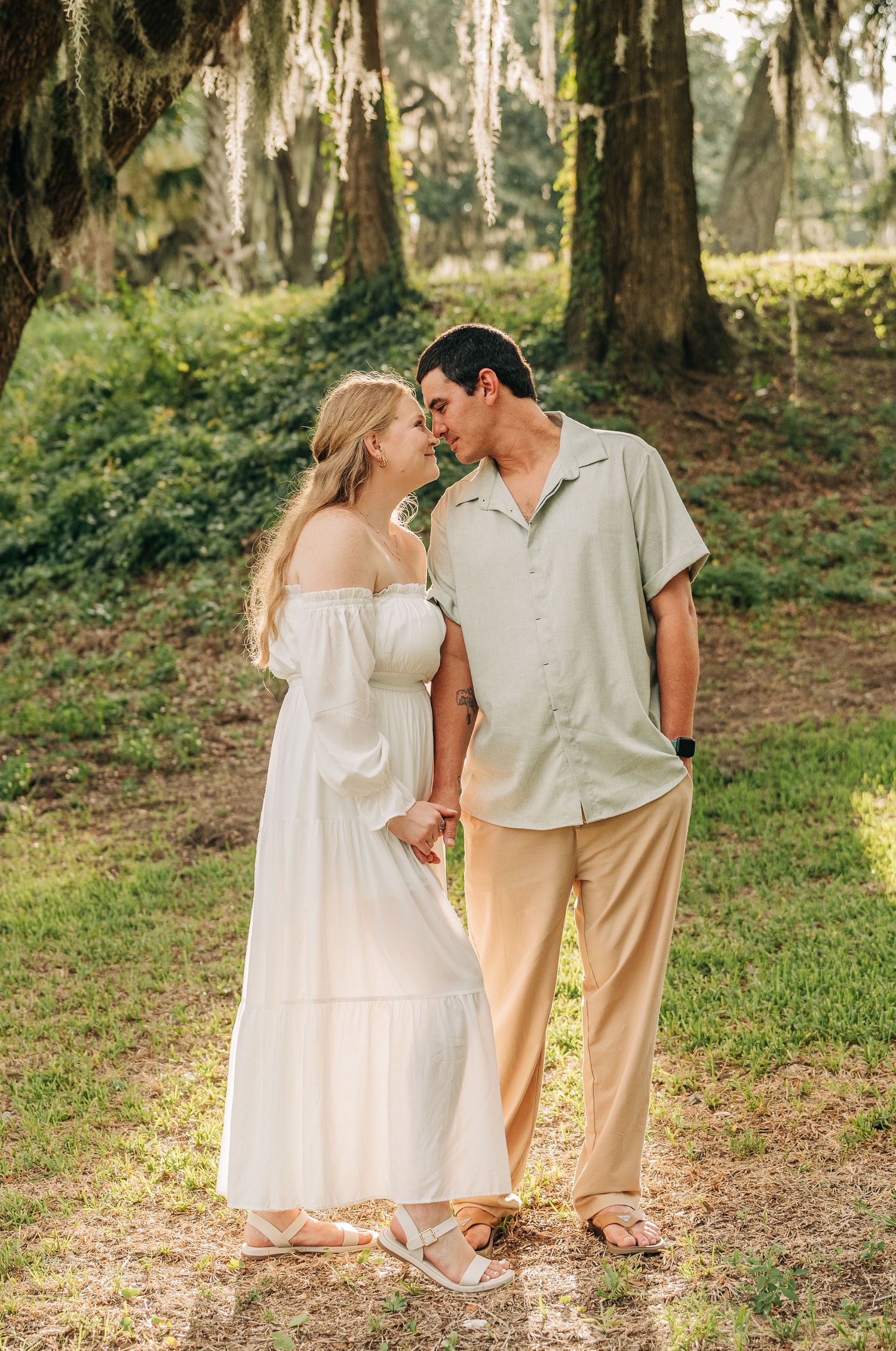 Engaged couple sharing a tender moment under oak trees draped in Spanish moss, the woman in a white dress.
