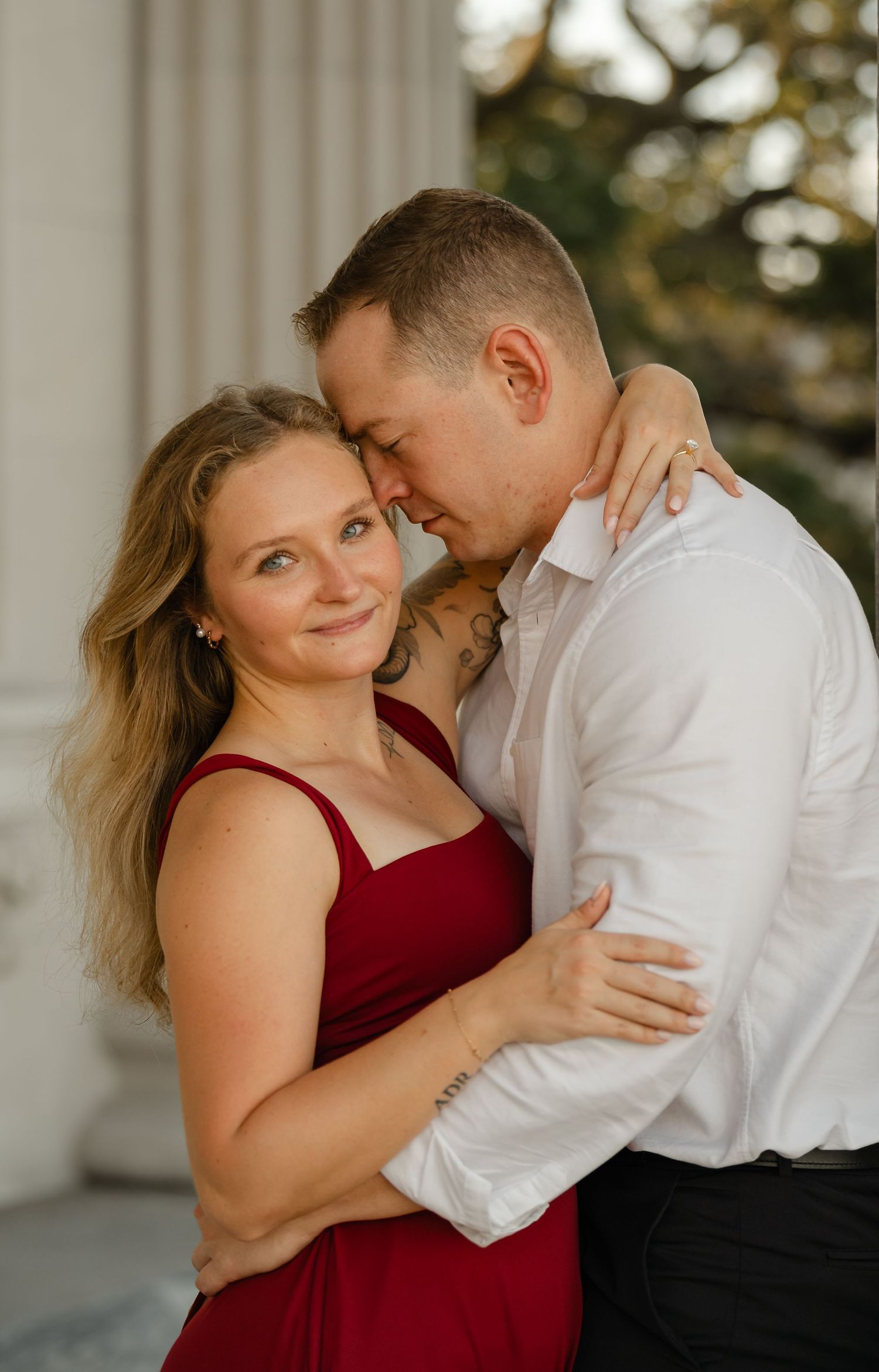 Close-up engagement portrait of a smiling woman in a red dress hugged by her fiancé in a white shirt near a stone column.