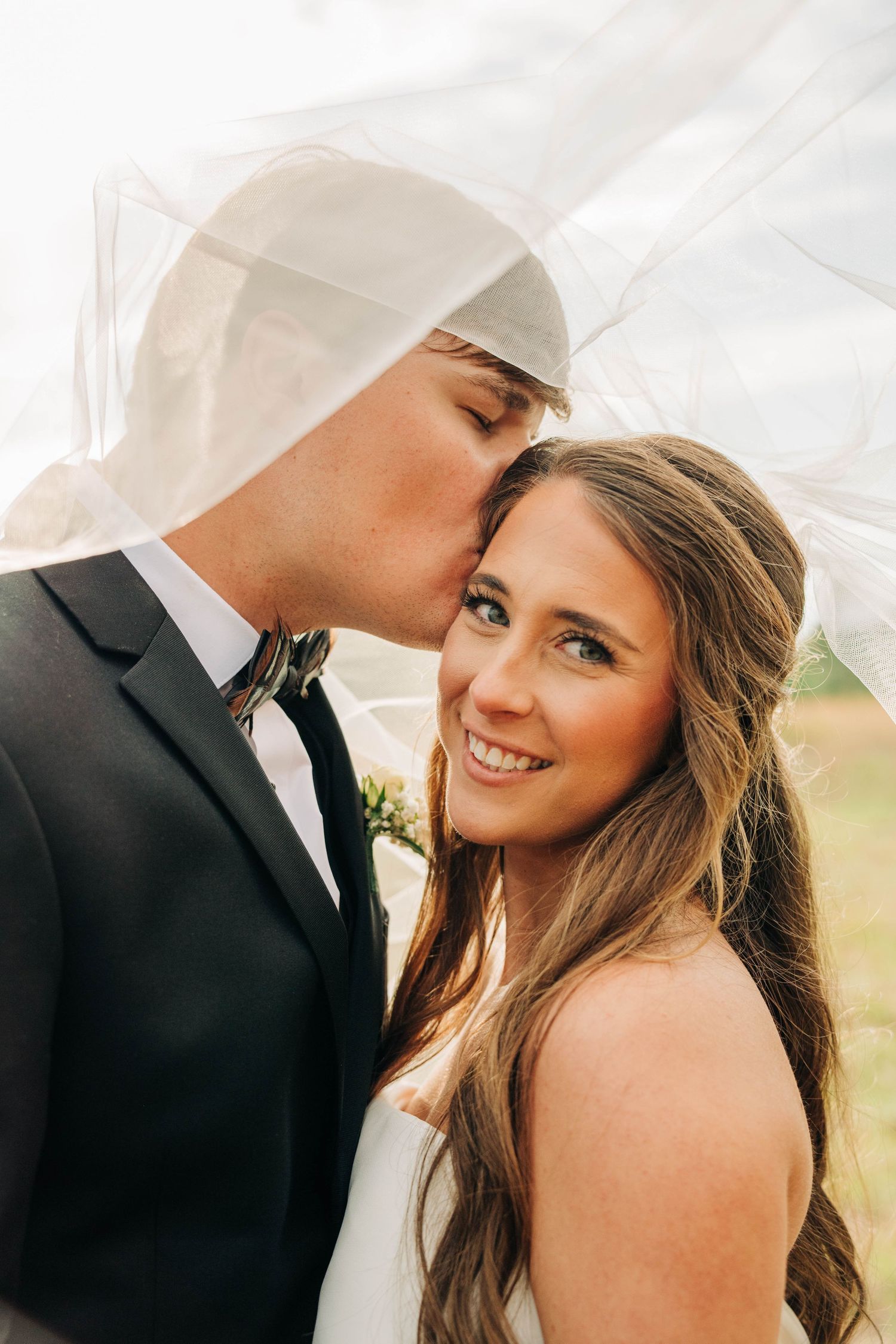 A groom kisses his smiling bride on the forehead beneath her sheer veil during an outdoor wedding portrait.