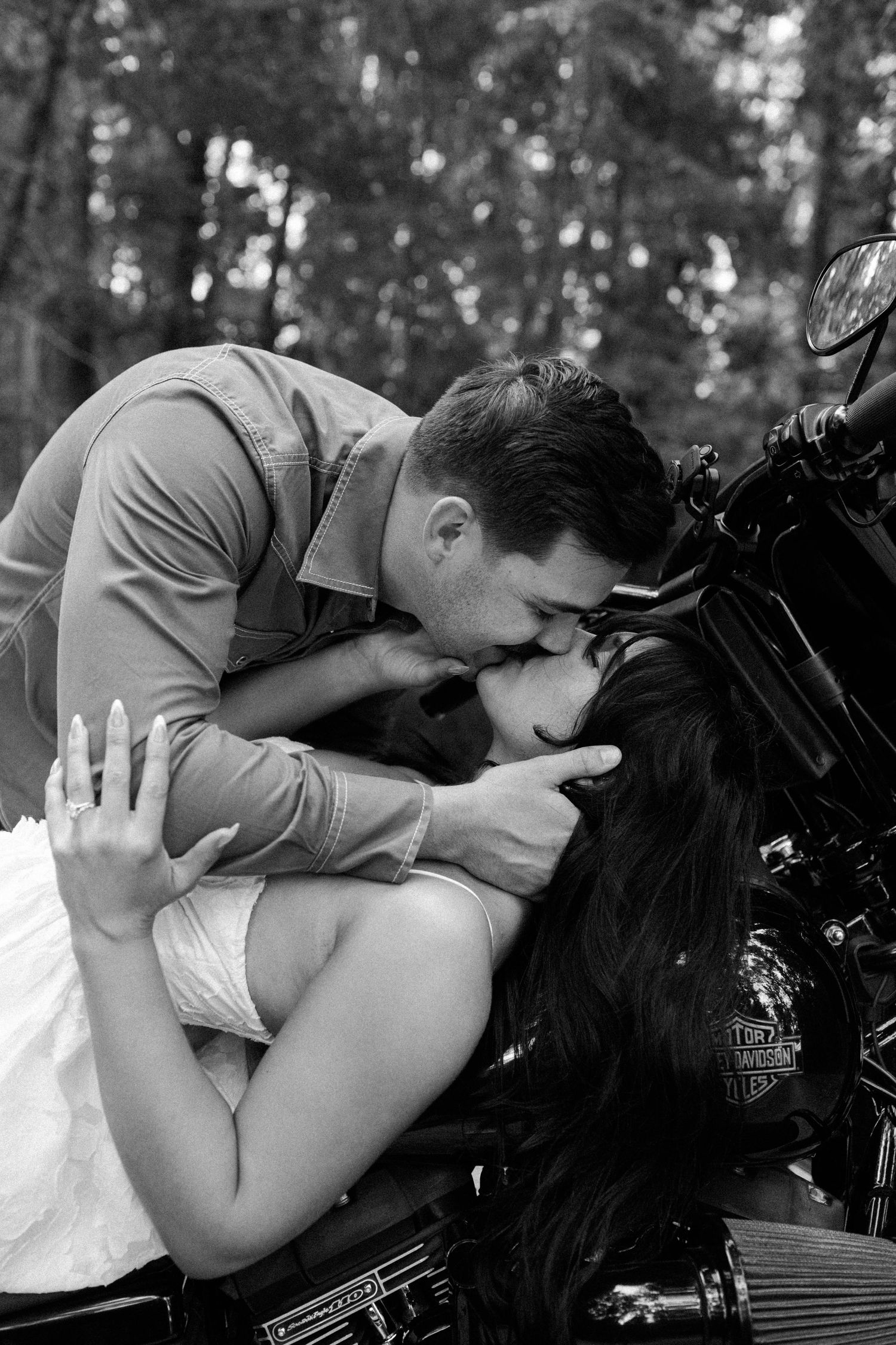 Black and white photo of a man leaning down to kiss a woman lying back on a Harley-Davidson motorcycle in the woods.