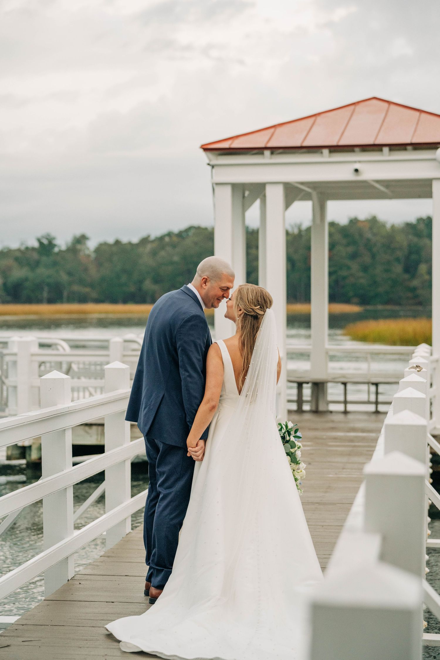 Bride and groom share a tender moment on a white wooden dock near a gazebo overlooking a marsh at their waterfront wedding.
