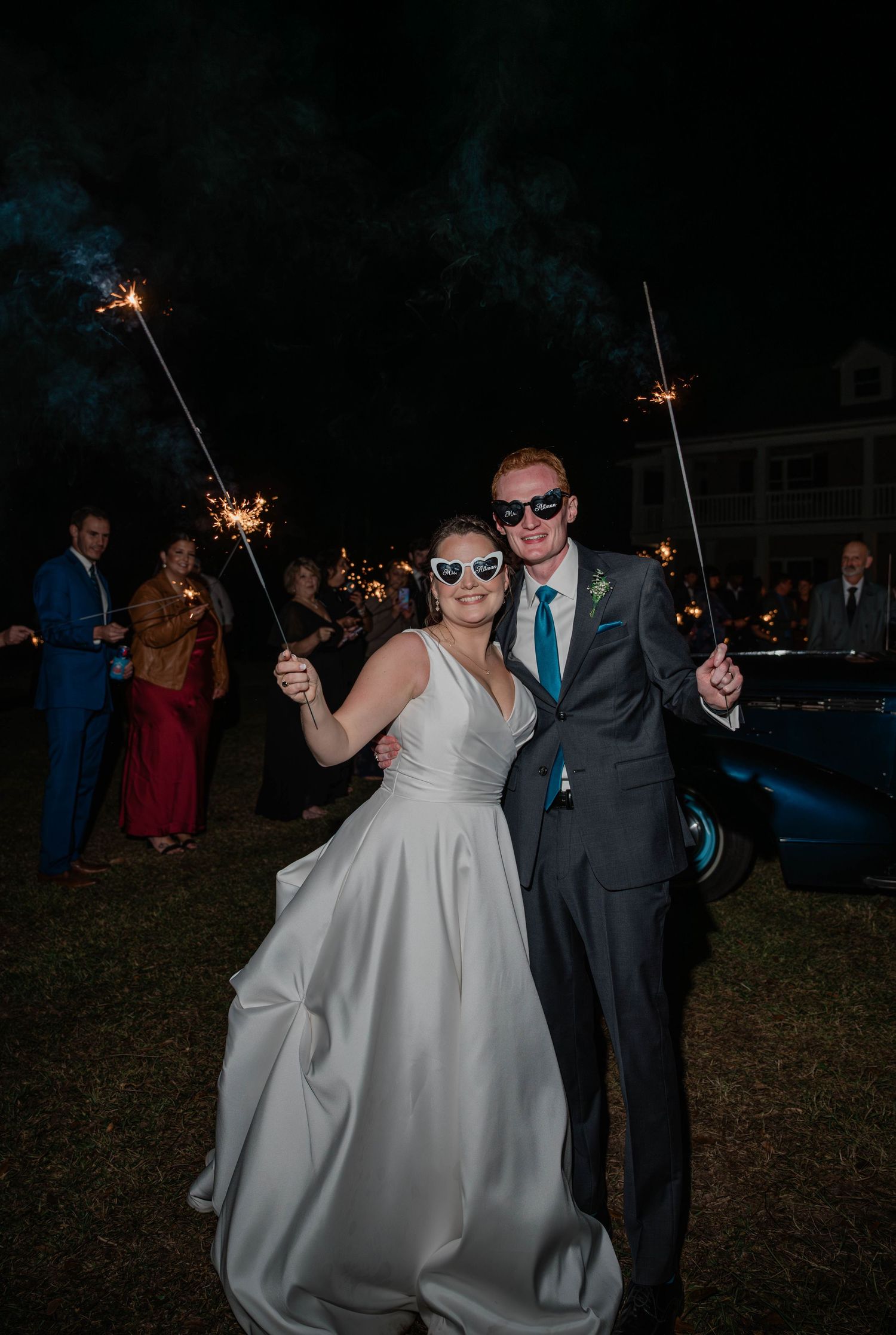 A smiling bride in heart-shaped sunglasses and a groom in sunglasses hold sparklers at night during their wedding send-off.