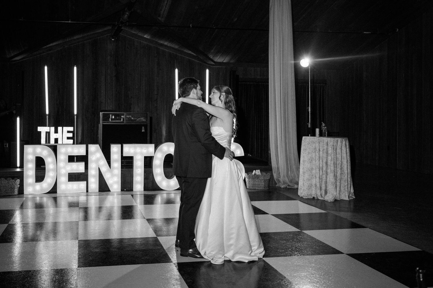 Bride and groom share a first dance on a checkered floor beneath large marquee letters spelling THE DENTON in black and white.