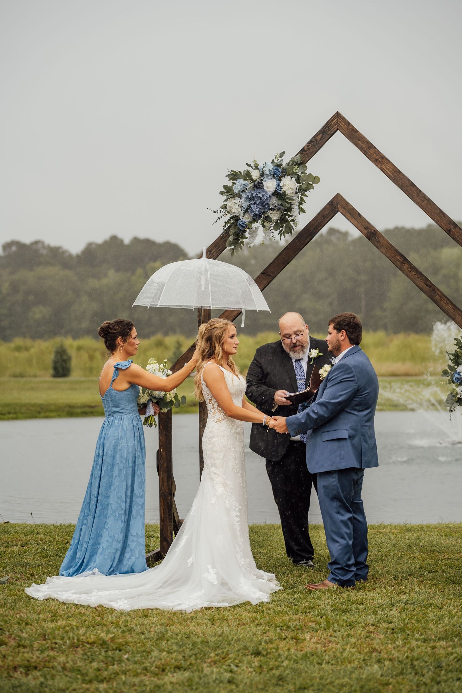 Bride and groom hold hands during an outdoor lakeside wedding ceremony under a geometric wooden arch as a bridesmaid holds an umbrella.