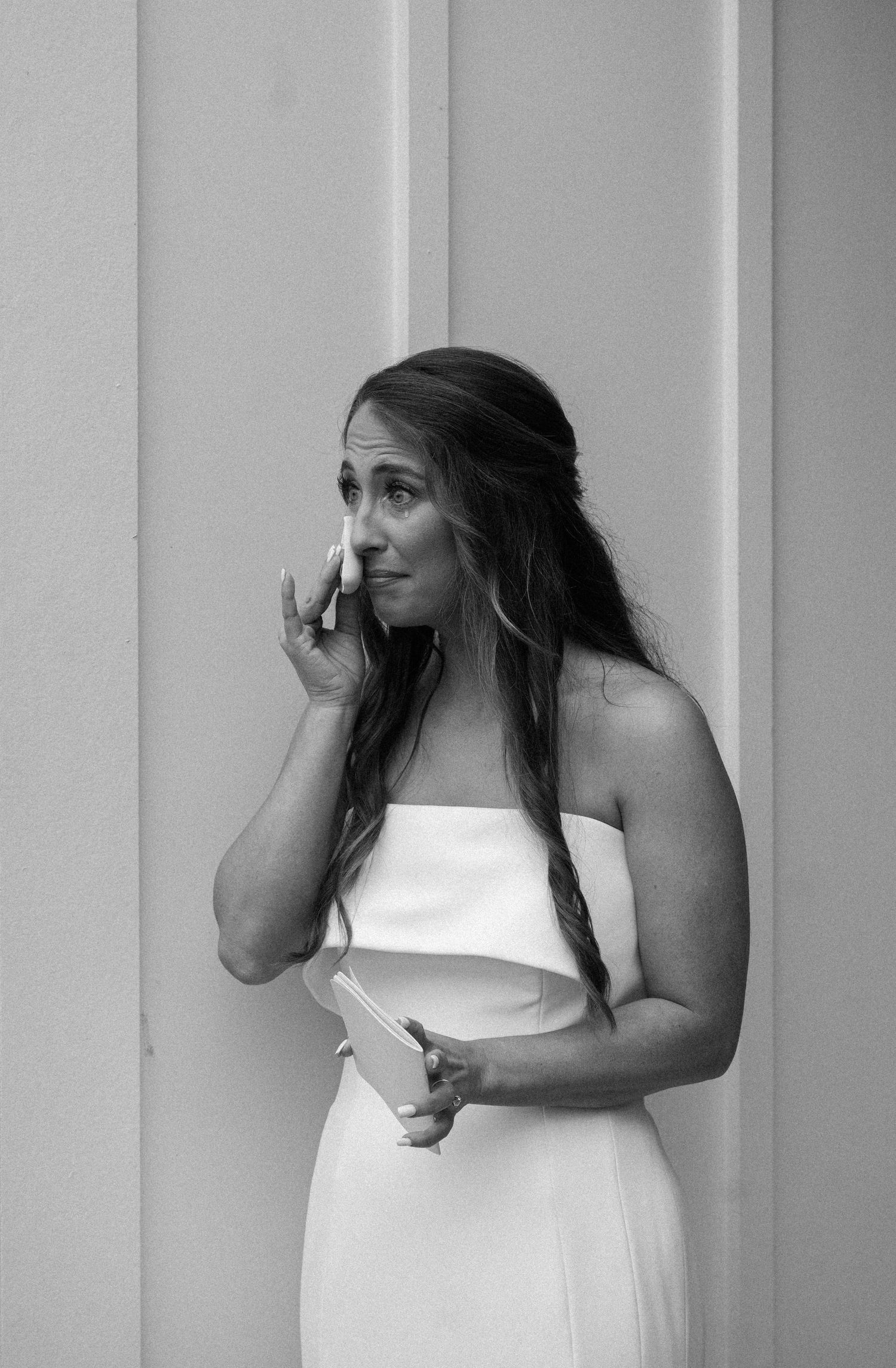 Black and white photo of an emotional bride in a strapless white gown wiping a tear while holding a small card.