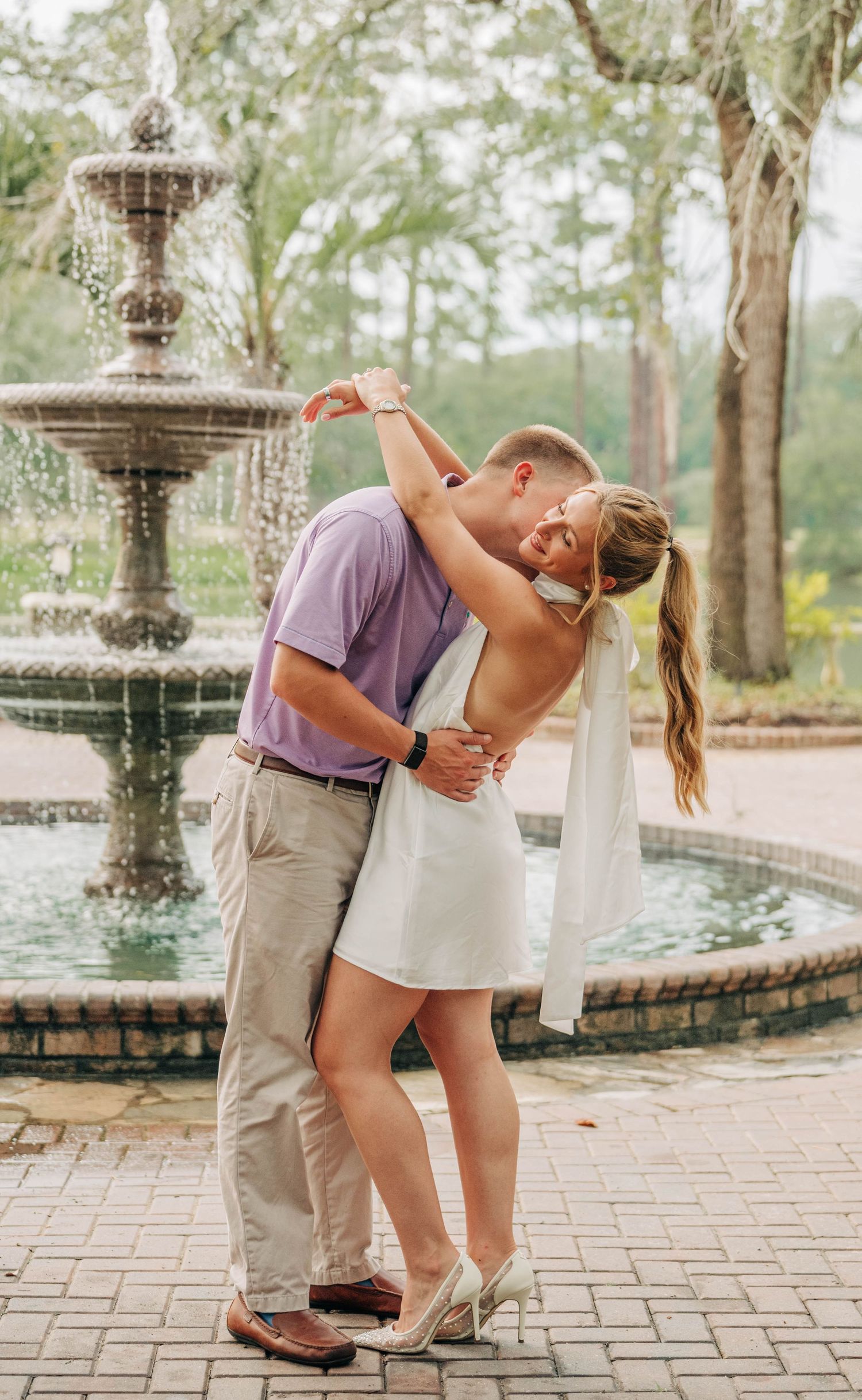 Couple in a lavender polo and white dress embrace and kiss beside a tiered stone fountain during an outdoor engagement photo session.