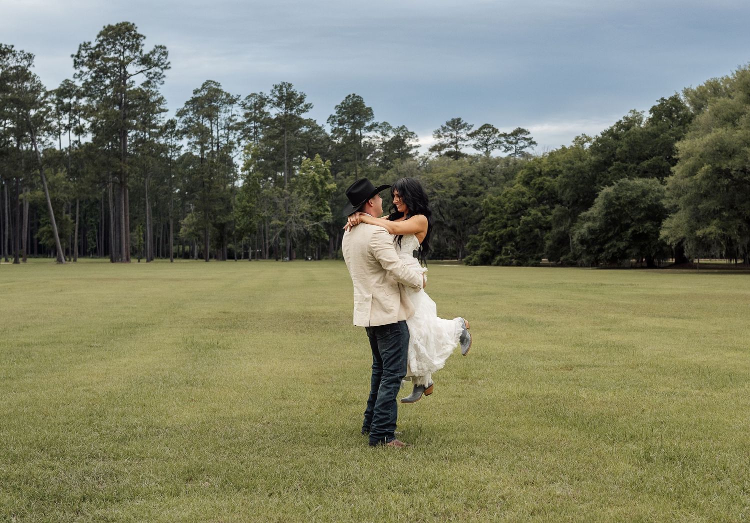 Groom in a cowboy hat lifts his bride in a white wedding dress in a grassy field with tall pine trees under a cloudy sky.