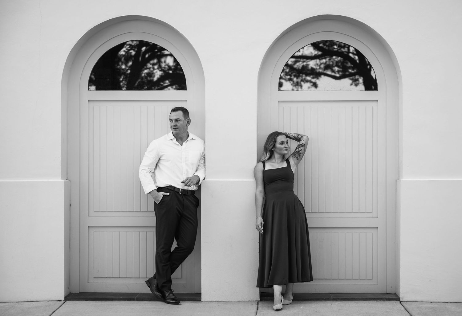 Black and white engagement photo of a couple leaning on separate arched doorways with tree-reflecting windows.
