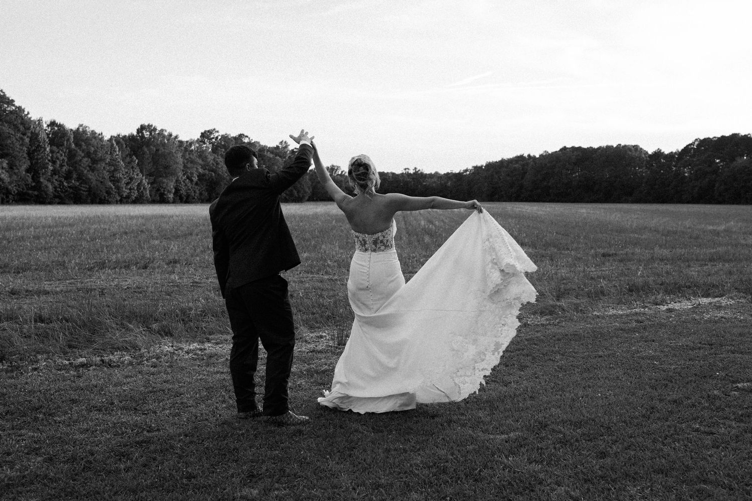 A black and white image of a bride and groom walking through an open field, her arm raised and her long gown flowing behind her.