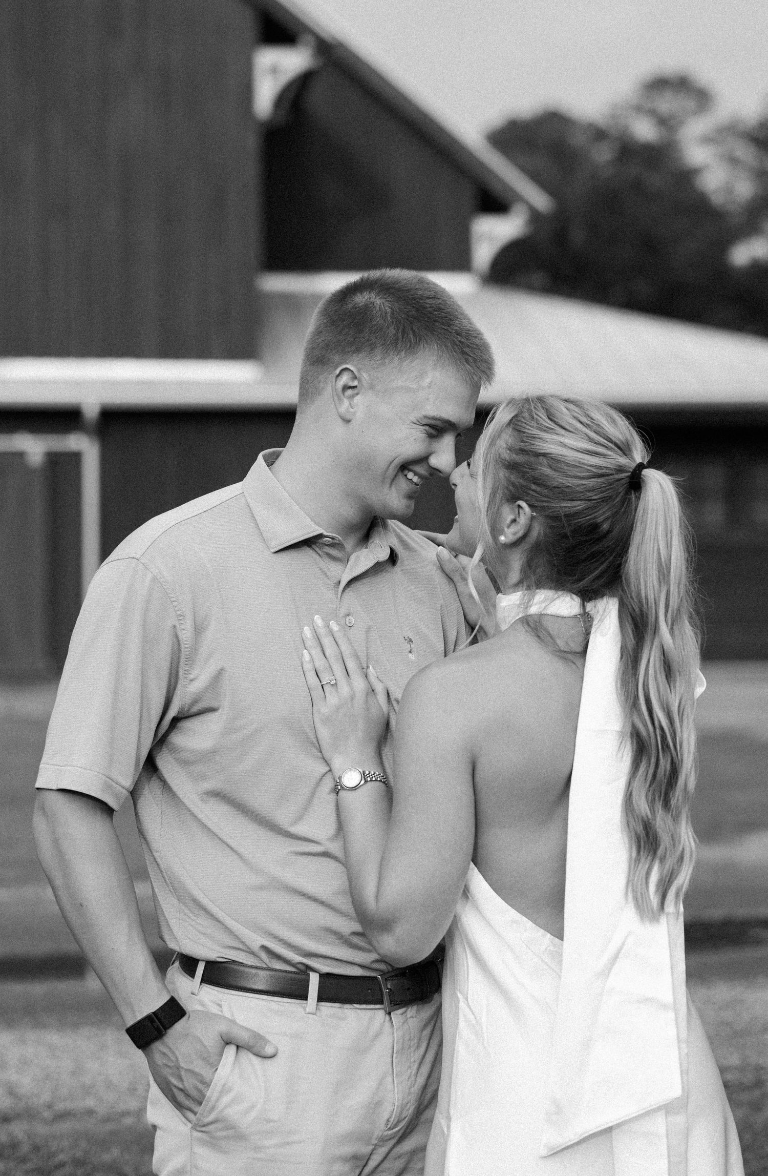 Black and white photo of a smiling couple nose to nose in front of a rustic barn, her hand resting on his chest.