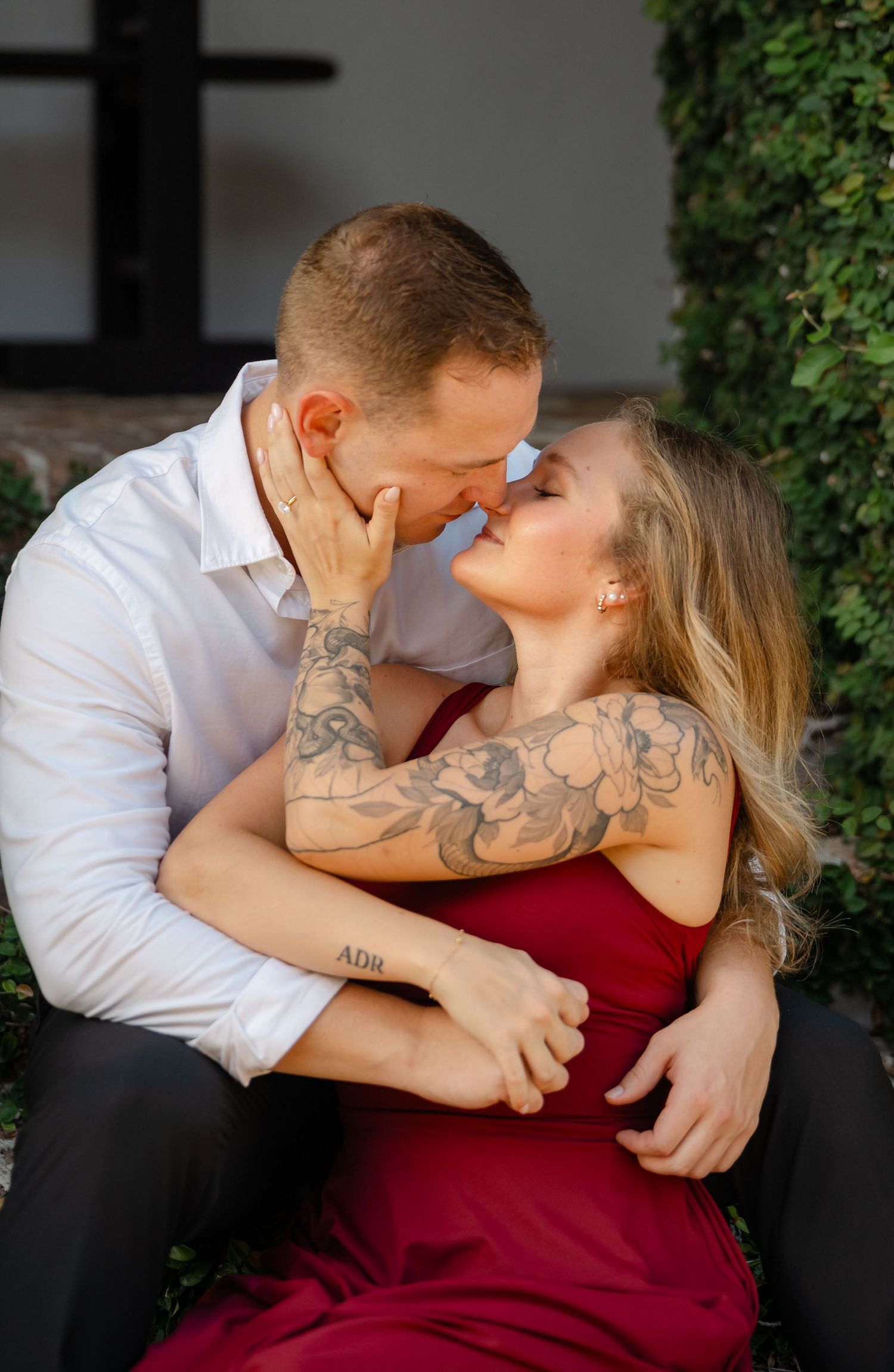 Couple in a close embrace nearly kiss during an engagement session, the woman in a red dress with floral arm tattoos.