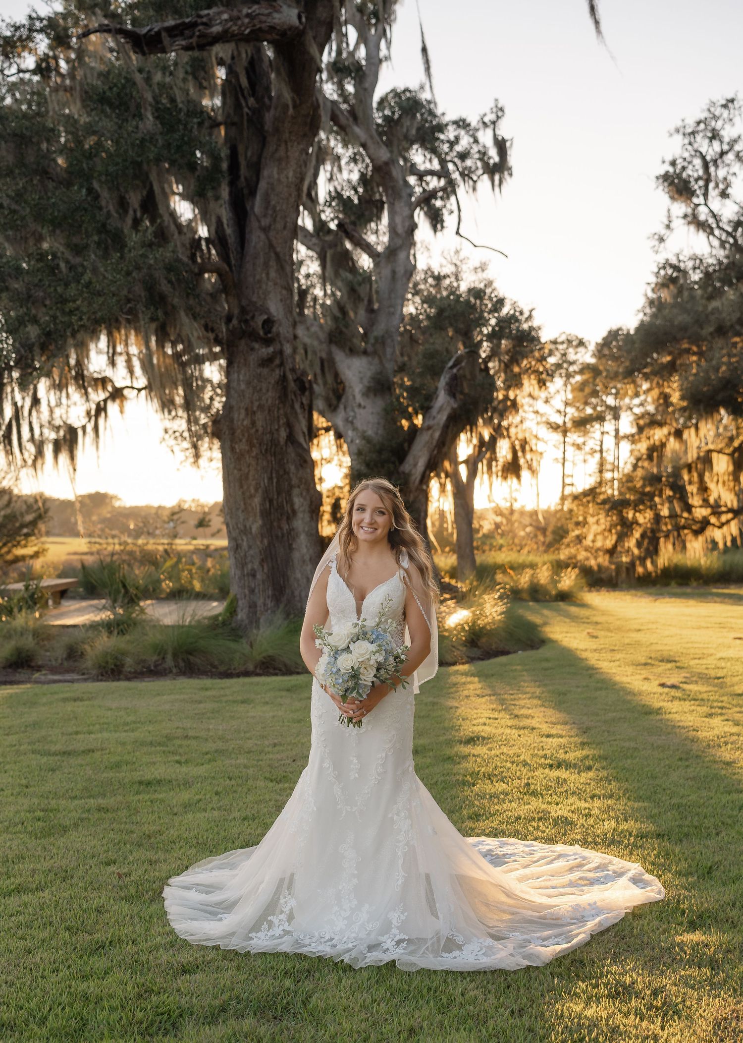 Bride in a lace fitted gown holds a white bouquet beneath moss-draped oak trees at golden hour sunset.