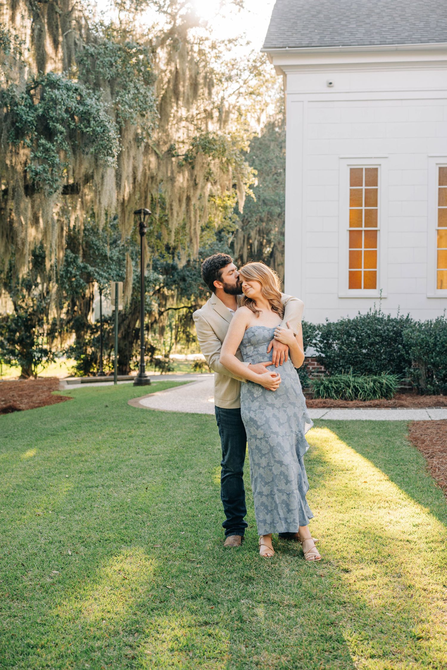 Couple embracing on a sunlit lawn beneath Spanish moss-covered oaks next to a white building at golden hour.