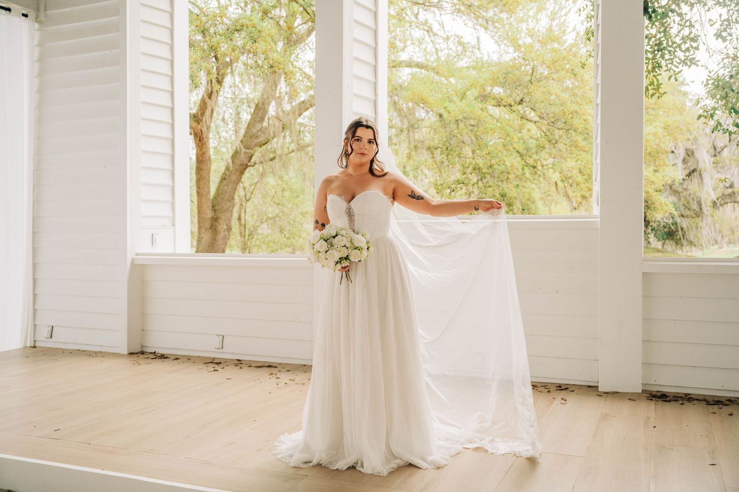 Bride in a soft tulle gown holds her veil out and a white bouquet in a bright pavilion with large windows.