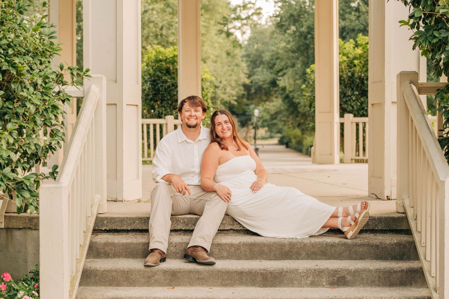 Smiling couple sits together on steps in front of a white gazebo surrounded by greenery during an engagement session.