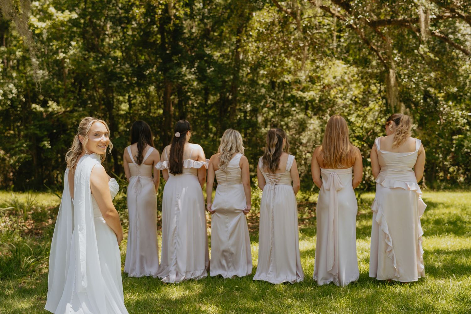 Bride faces six bridesmaids in matching light dresses standing in a row on a sunny grassy lawn framed by trees.