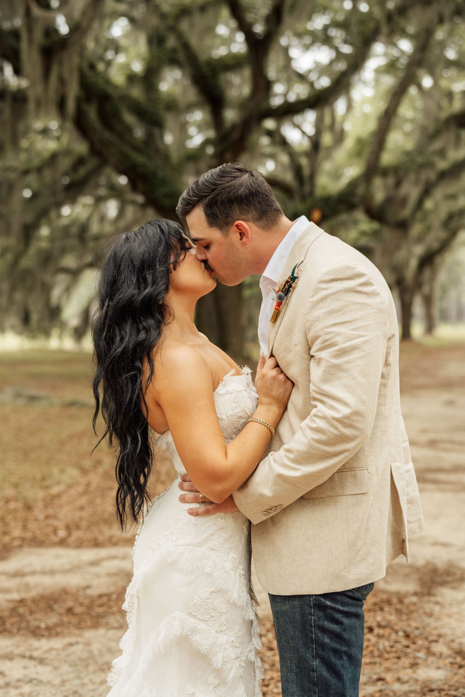 Bride and groom in a tan blazer share a romantic kiss outdoors beneath live oak trees draped in Spanish moss.