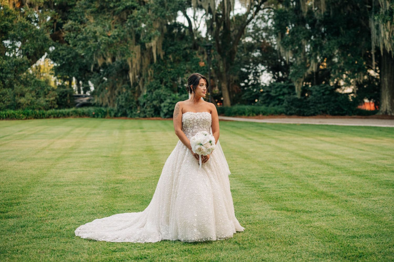 Bride in a strapless beaded wedding gown with a long train holding a bouquet on a manicured lawn beneath moss-draped oak trees.